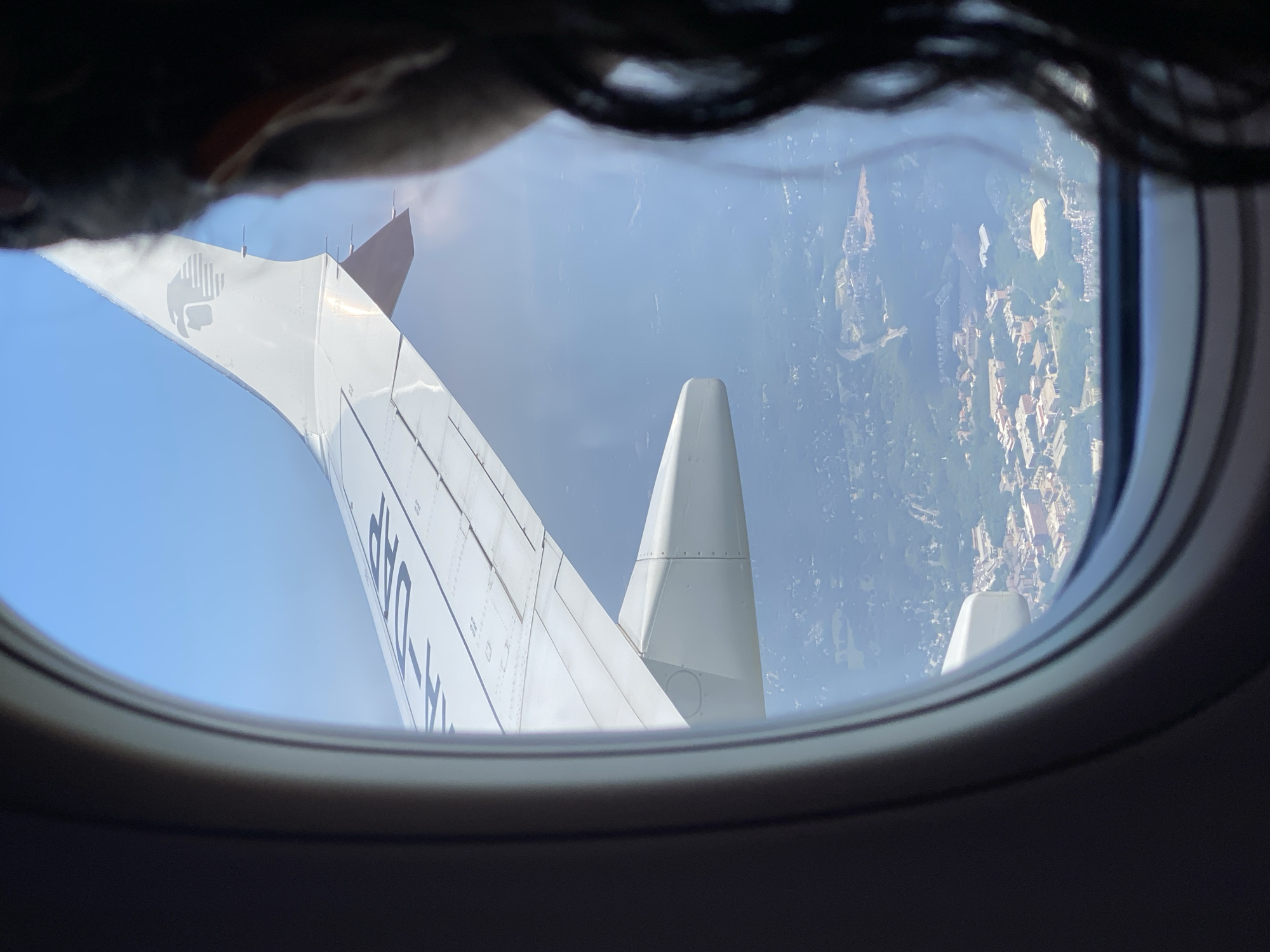 The image is taken from an airplane window, looking out at the landscape below. A person's dark hair is visible in the foreground, obscuring part of the view. The airplane's wing, with the Aeromexico logo and part of the aircraft registration "XA-DAP" visible, dominates the upper portion of the frame. Below the wing, a suburban area is visible, with buildings, trees, and roads. The sky is clear and blue. Based on the visible landmarks and the distinct architecture of the buildings in the area, this location is likely the campus of Rowan University in Washington Township, New Jersey, United States of America. The oval stadium in the center of the campus is a recognizable feature.