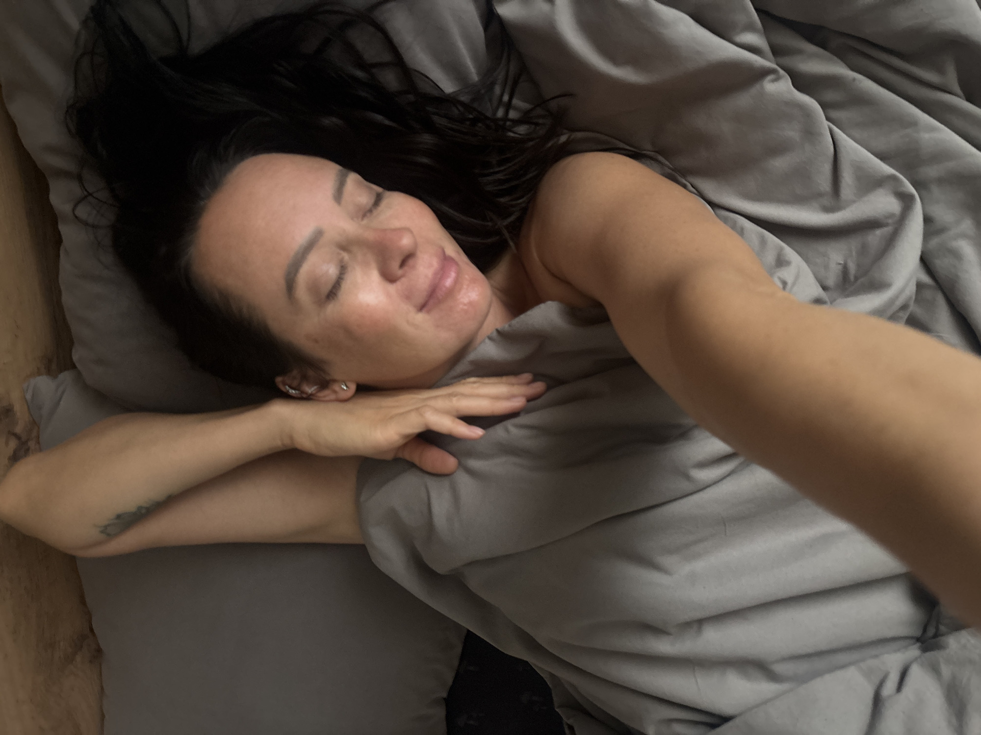 A woman with dark hair lies in a bed with gray bedding. Her eyes are closed, and she appears to be resting. Her right arm is extended towards the camera in a selfie-style pose. A tattoo is visible on her left arm. The location cannot be determined from the image.