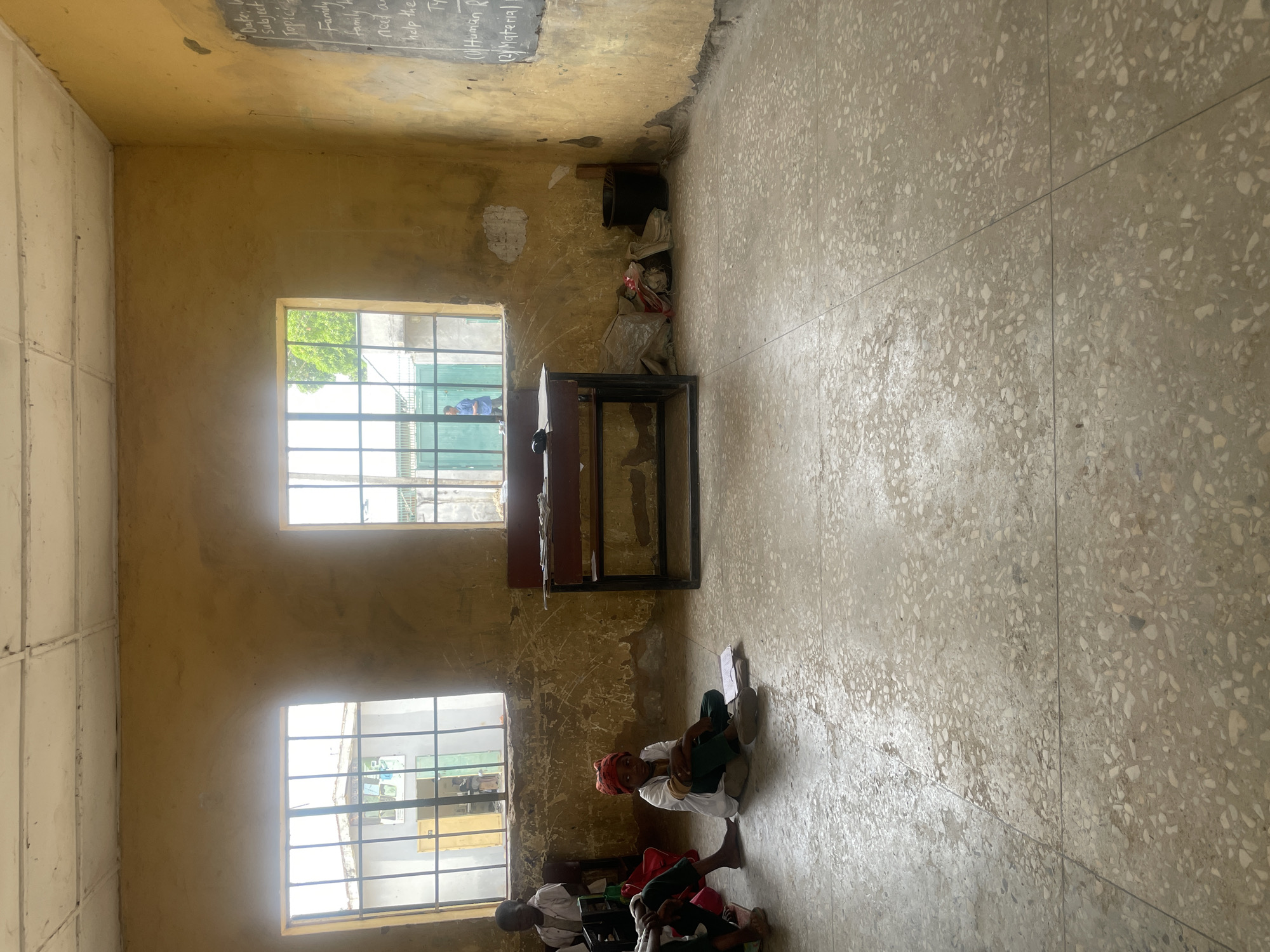 The image depicts an indoor scene, likely a classroom, in Unguwar Rimi, Nigeria. The room has yellow walls with two windows. A chalkboard with handwriting is visible in the upper left corner. A desk stands against the wall between the windows. On the floor, several people are sitting. One individual is wearing a red head covering and a white shirt. Debris and a trash receptacle can be seen in the back corner of the room. The floor is made of grey tile.