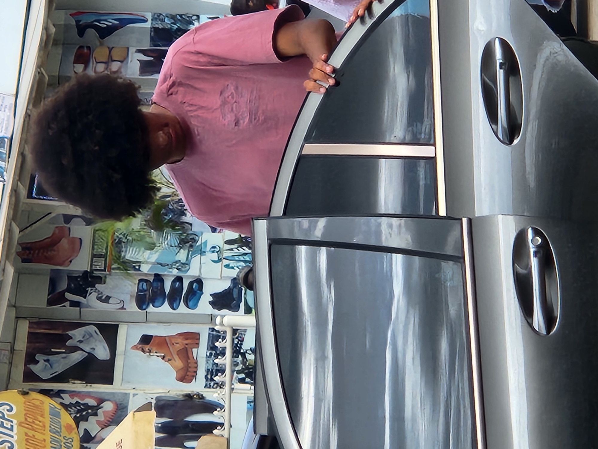 A young Black male with a large afro and wearing a pink t-shirt stands by the open passenger door of a dark gray sedan. He is reaching into the car with his right hand. Behind him, the wall of a structure displays various images of footwear, suggesting a shoe repair shop. The environment appears to be outdoors, possibly on a street in Jos, Nigeria. The overall lighting suggests it is daytime.