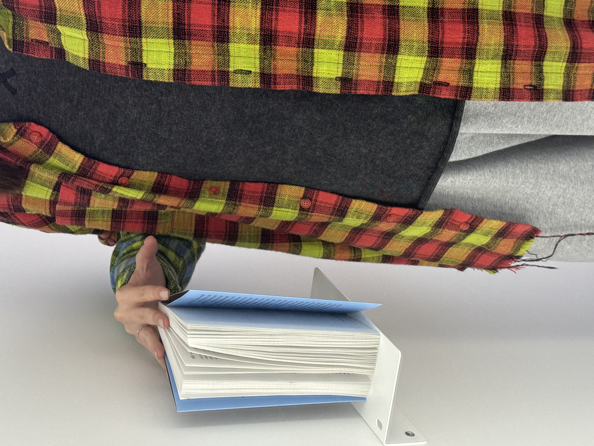 The image shows a person holding a book in a white metal book holder. The book has a light blue cover. The person is wearing a red, yellow, and orange plaid shirt, a dark gray shirt underneath, and light gray sweatpants. The background is a plain white surface. The location is Unknown, Unknown.