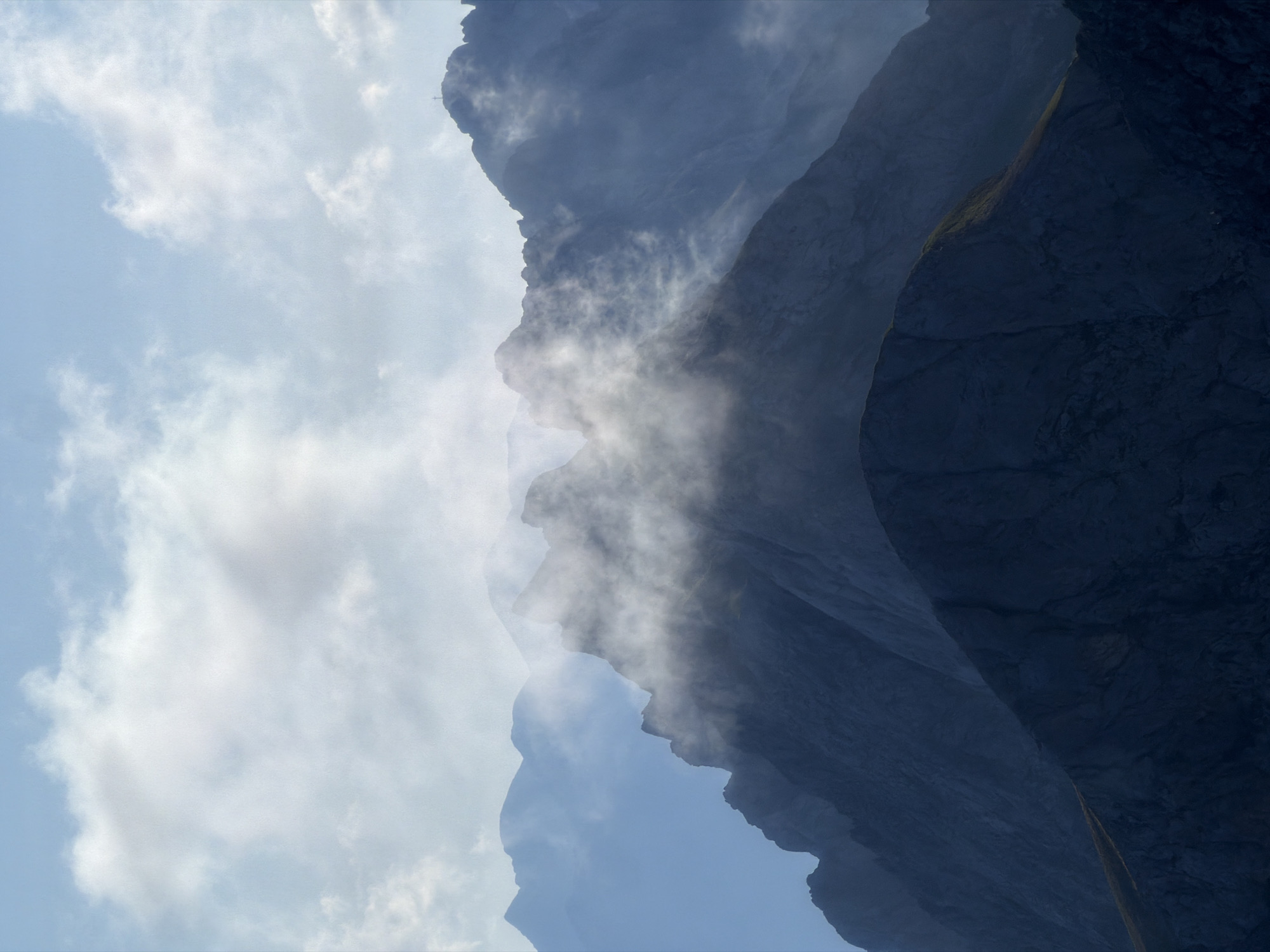 The image depicts a serene mountain landscape. The foreground features dark, rugged mountain peaks shrouded in deep shadow, with a hint of illuminated grass on the left slope of the foremost peak. As the eye moves towards the background, layers of progressively lighter, hazy mountains recede into the distance. Wisps of white mist weave through the middle ground, obscuring some of the mountain ridges and adding a sense of depth and mystery to the scene. The sky above is a pale blue, filled with scattered, soft white clouds, some of which are illuminated by diffused sunlight. The overall impression is one of a tranquil, majestic natural environment. This scene could be located in the vicinity of Grainau, Germany, a region known for its stunning alpine scenery, particularly the Zugspitze massif which features similar dramatic mountain formations and potential for mist and cloud cover. There are no people or identifiable man-made structures visible in the image.