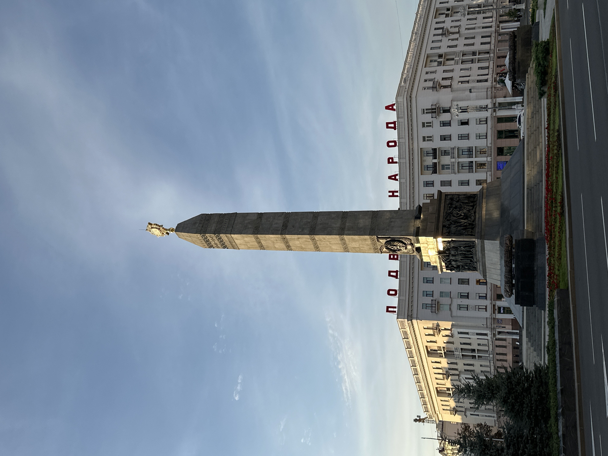 This is a full, eye-level shot of the Victory Monument in Minsk, Belarus. The monument, a tall obelisk topped with a golden star and eagle, stands in the center of a plaza. The monument is flanked by a long, multi-story white building with numerous windows and balconies. Red lettering on the building reads "ПОБЕДЫ НАРОДА" (Victory of the People). In the foreground, a street with traffic lanes and a median is visible. Evergreen trees and flowerbeds are also present around the monument. The sky is blue with scattered clouds. There are no people visible in the image.