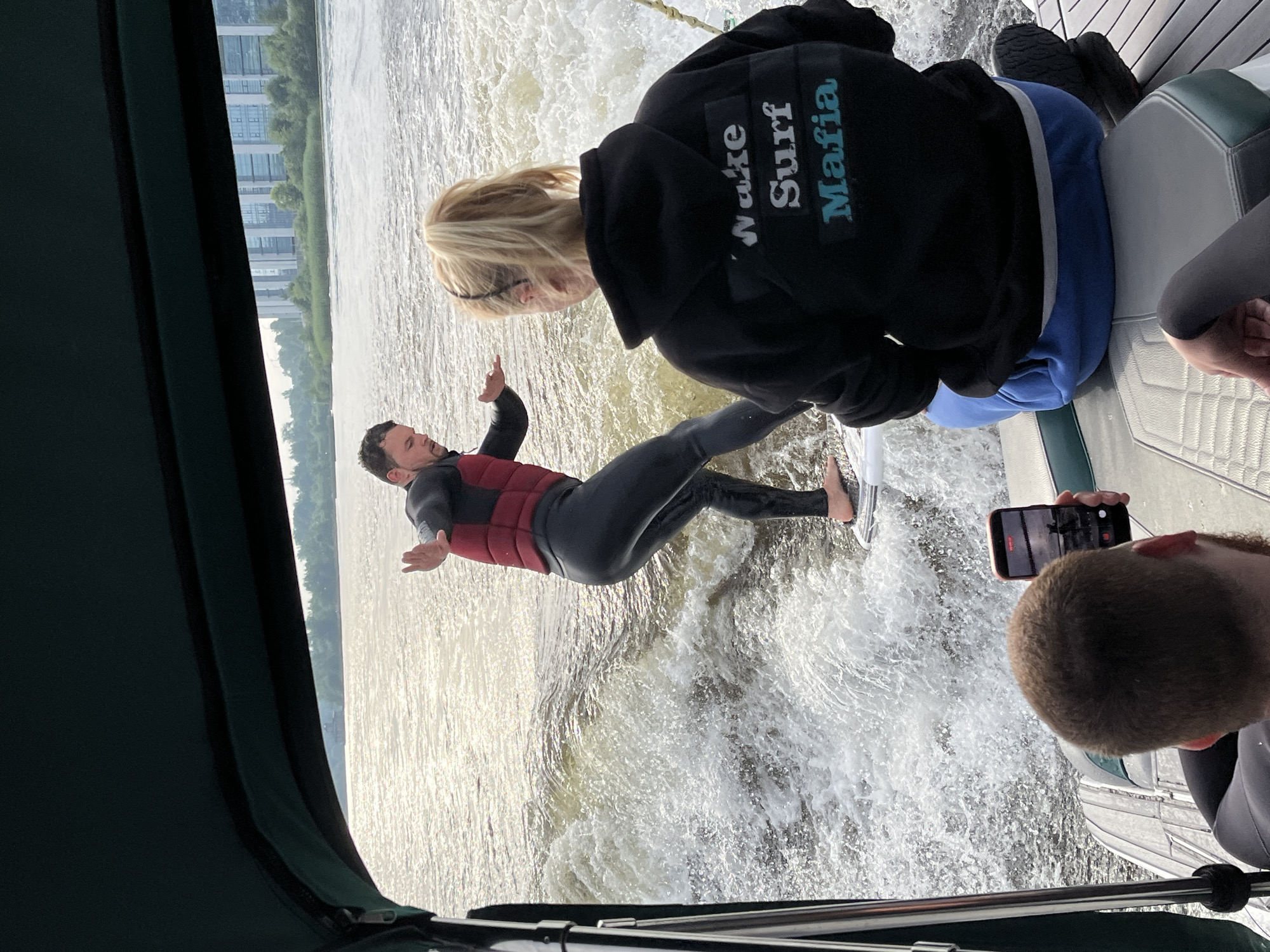 In Saint Petersburg, Russia, on a bright, sunny day, three individuals are on a boat. A man in a black wetsuit and a red vest is wake surfing behind the boat, his arms outstretched for balance on a white surfboard. In the boat, a woman with blonde hair in a ponytail wears a black hoodie with the words "Wake Surf Mafia" in white and aqua lettering. Another person, only their back and head visible, is seated in the boat, holding a smartphone to record the wake surfer. The boat is on a body of water, possibly a river or bay, with a city skyline visible in the distance, featuring modern buildings and trees. The water is choppy and creating a significant wake.