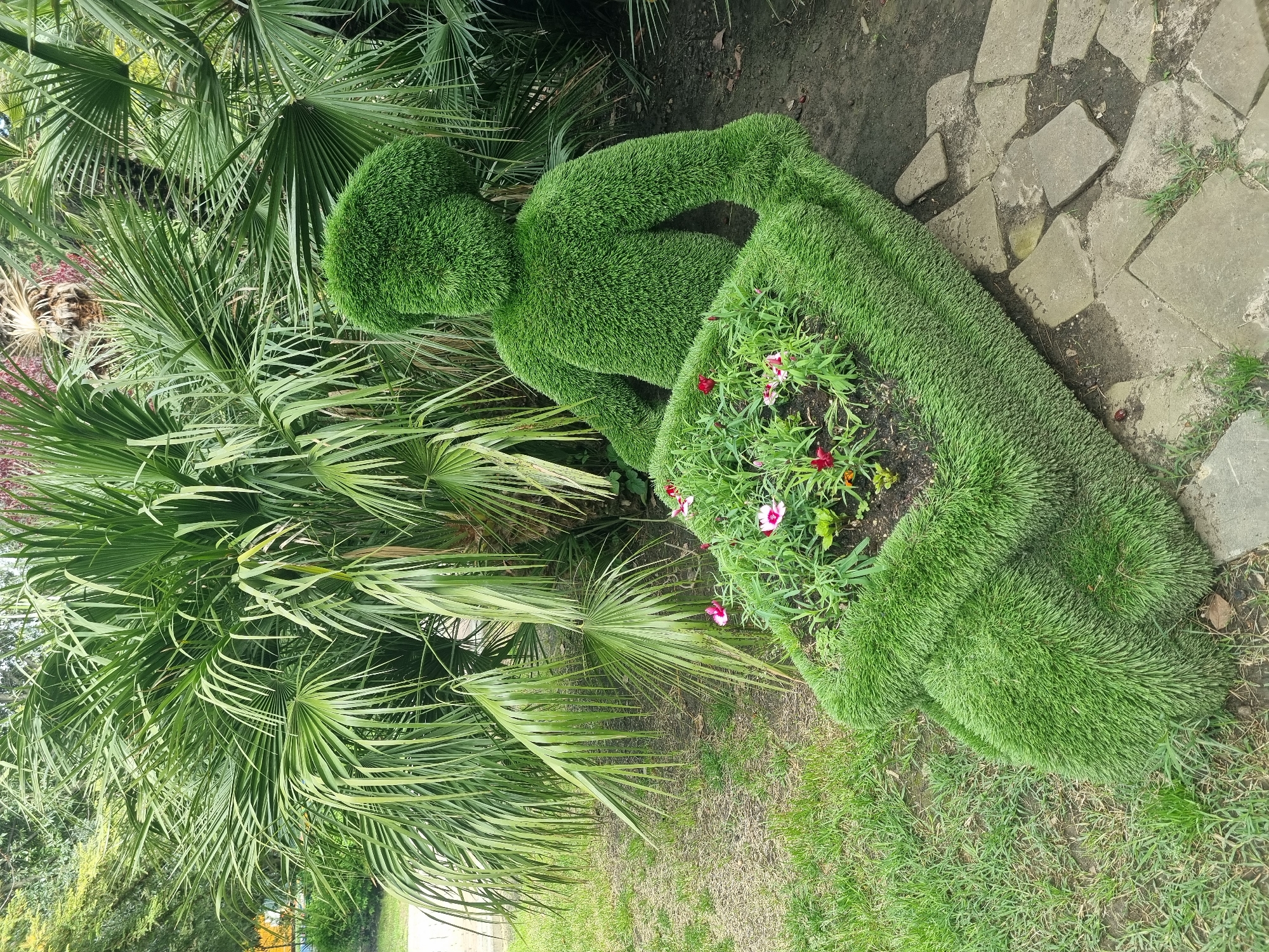 The image shows a topiary in the city of Sochi, Russia. The topiary is in the shape of a person with a wheelbarrow filled with flowers. The scene is set in a garden or park, with palm trees and stone pathways visible in the background.