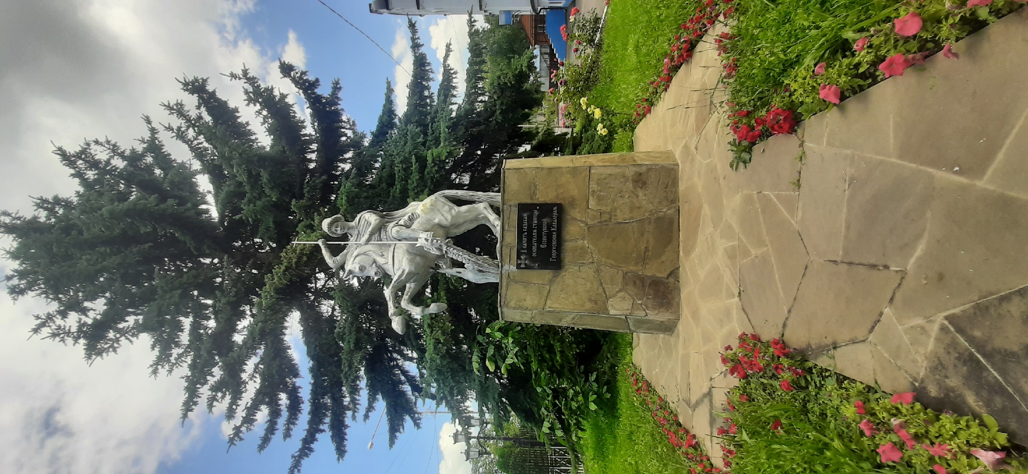 This is a frontal, eye-level, medium shot of a statue of Saint George slaying a dragon, with a large pine tree behind it. The statue is made of silver-colored metal and is mounted on a stone pedestal. A plaque on the pedestal reads, "In memory of the Cossack founders of the village of Yessentukskaya, Georgievsky Kavalery." The statue and pedestal are surrounded by a paved walkway and flowerbeds filled with red and pink flowers. The sky is blue with scattered clouds, and the overall lighting suggests it is daytime.