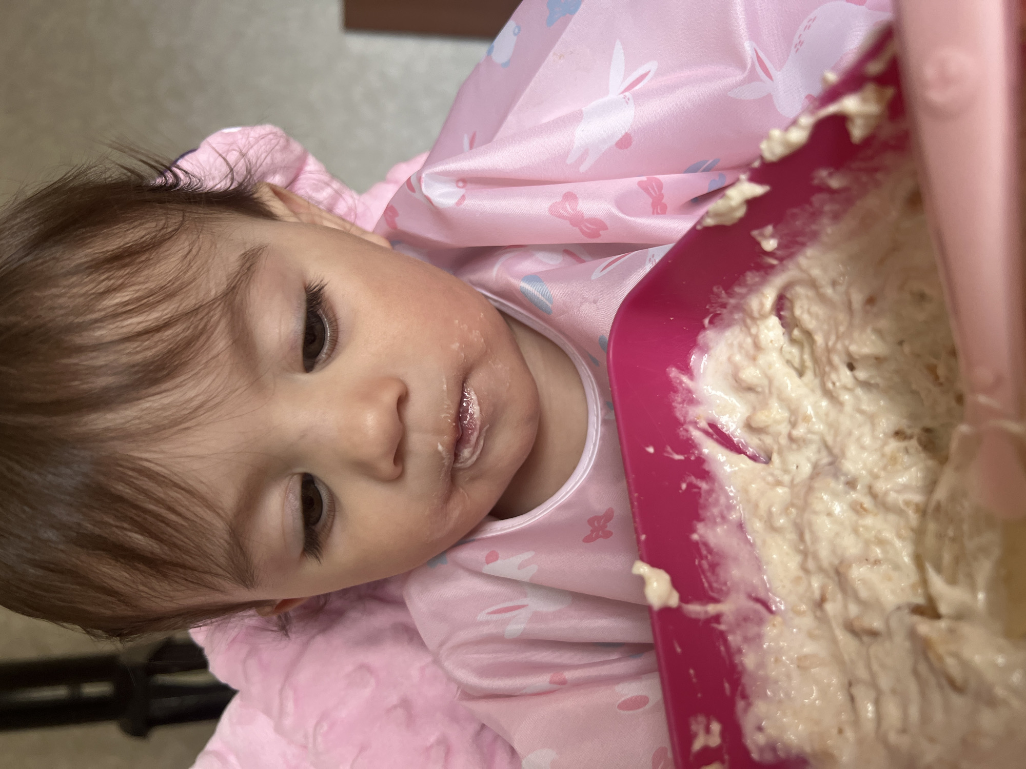 The image displays a close-up, slightly tilted view of a baby or toddler during mealtime, likely in an indoor setting in Elena, Bulgaria. The central subject is a child, appearing to be a girl, with dark brown hair, long eyelashes, and brown eyes. Her face shows evidence of eating, with light-colored food residue around her mouth and on her left cheek.
The child is dressed in a pink long-sleeved garment, possibly a bib or top, featuring patterns of white and light blue bunnies and bows. She is positioned against a soft, light pink blanket or cushion. To her right, a bright pink bowl contains a substantial amount of thick, light-colored food, which appears to be oatmeal or a similar pureed meal, showing distinct textures. A matching pink spoon is visible within the food, suggesting active eating.
The baby's expression is neutral, without strong indications of emotion. The background is softly out of focus, providing no specific details about the time of day, weather, or other location cues beyond the provided geographical context of Elena, Bulgaria. There is no visible text in the image.