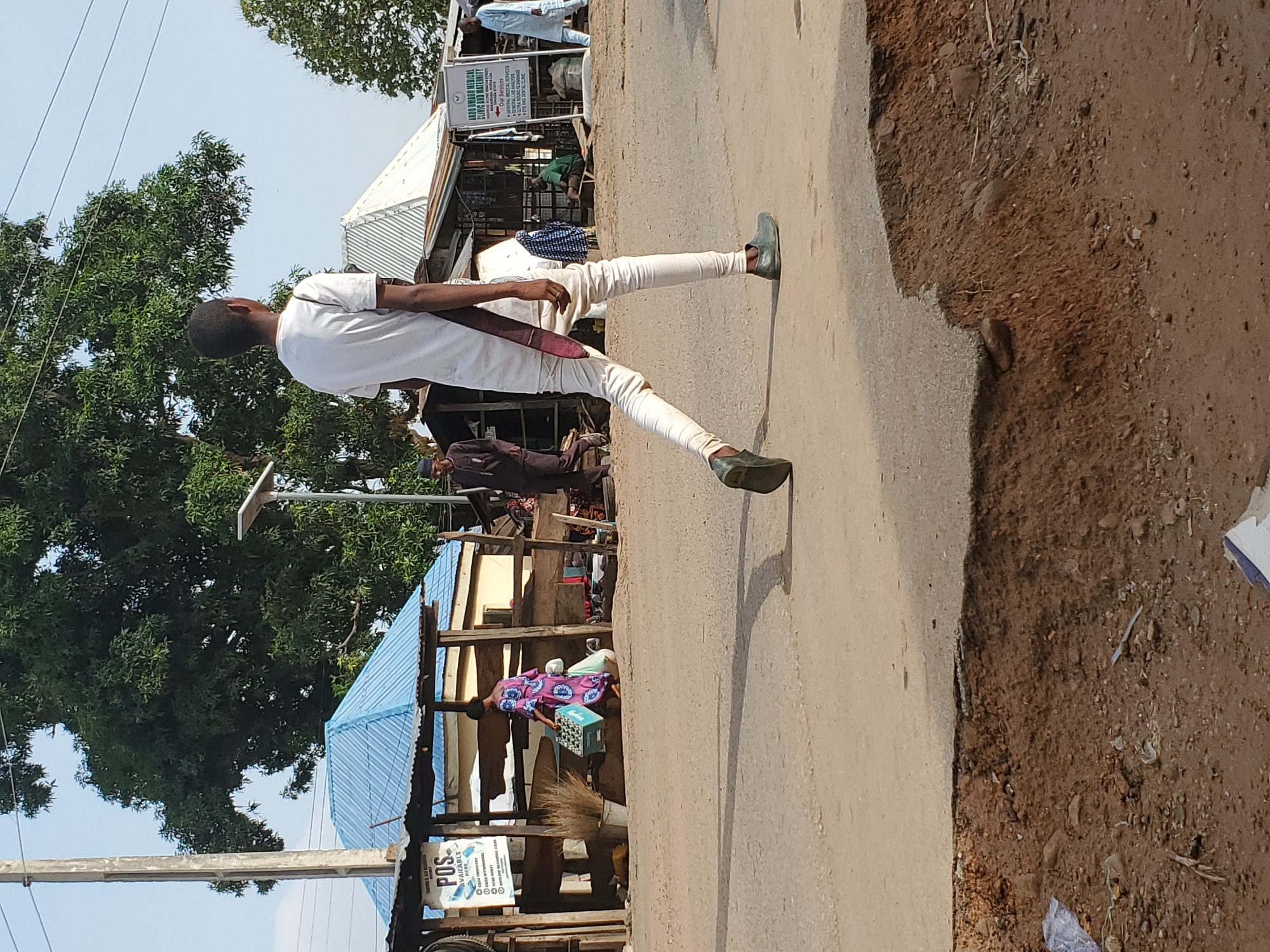 The image captures a lively street scene in Mararaba, Nigeria, under bright daylight. The main subject is a young man, viewed from behind, walking on a light-colored paved road or sidewalk. He wears a white short-sleeved top, form-fitting white trousers, and dark-colored shoes or sandals, with a dark strap or bag slung across his body.
To the left of the man, behind him, are various makeshift structures resembling market stalls or small shops with corrugated metal roofs, some blue and some silver. Lush green trees flank the background on the left. Several people are visible in the midground and background, some appearing to be vendors or customers near the stalls, while others walk along the road. One person in a patterned dress is seen near a blue crate and a broom.
Prominent signage is visible: one sign clearly reads "P.O.S AVAILABLE HERE." Another larger sign, situated above a shop, identifies it as "A.M.A UMARU MEDICAL CLINIC AND MATERNITY," listing services such as "MEDICAL X-RAY," "PHARMACY," "LABORATORY," "ANTENATAL," "GENERAL MEDICAL SERVICES," "FAMILY PLANNING," "ULTRASOUND SCANNING," and "EYE AND DENTAL CLINIC."
The paved road contrasts with a dusty, reddish-brown dirt shoulder or path to the right. Shadows suggest a sunny day, likely around midday or early afternoon. The overall setting depicts a functional, bustling area with commercial activity alongside a main thoroughfare.