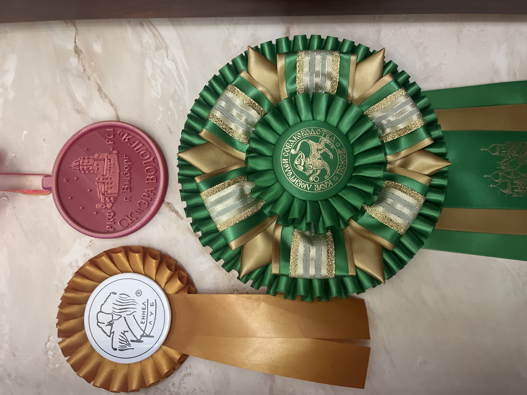 Three awards are displayed on a marble-patterned surface. On the left is a circular gold rosette with a white center featuring a black line drawing of a dog and the text "КЕННЕЛ КЛУБ". Two long gold ribbons hang from the rosette. In the upper center is a pink circular medal with a raised image of a building and text that appears to be in Russian, including "г. Выборг" and "FCI". Below these, to the right, is a large green rosette with gold accents and several smaller green and gold ribbons hanging from it. The center of this rosette contains a circular emblem with gold text and a gold illustration of a dog. One of the hanging ribbons has a gold illustration of what appears to be Orthodox church domes. The awards are arranged on a textured surface that resembles marble. The image is taken from a top-down perspective. There are no people or animals present. The scene appears to be an indoor display of awards. The location in Vladimirovka, Russia, is not discernible from the image itself.