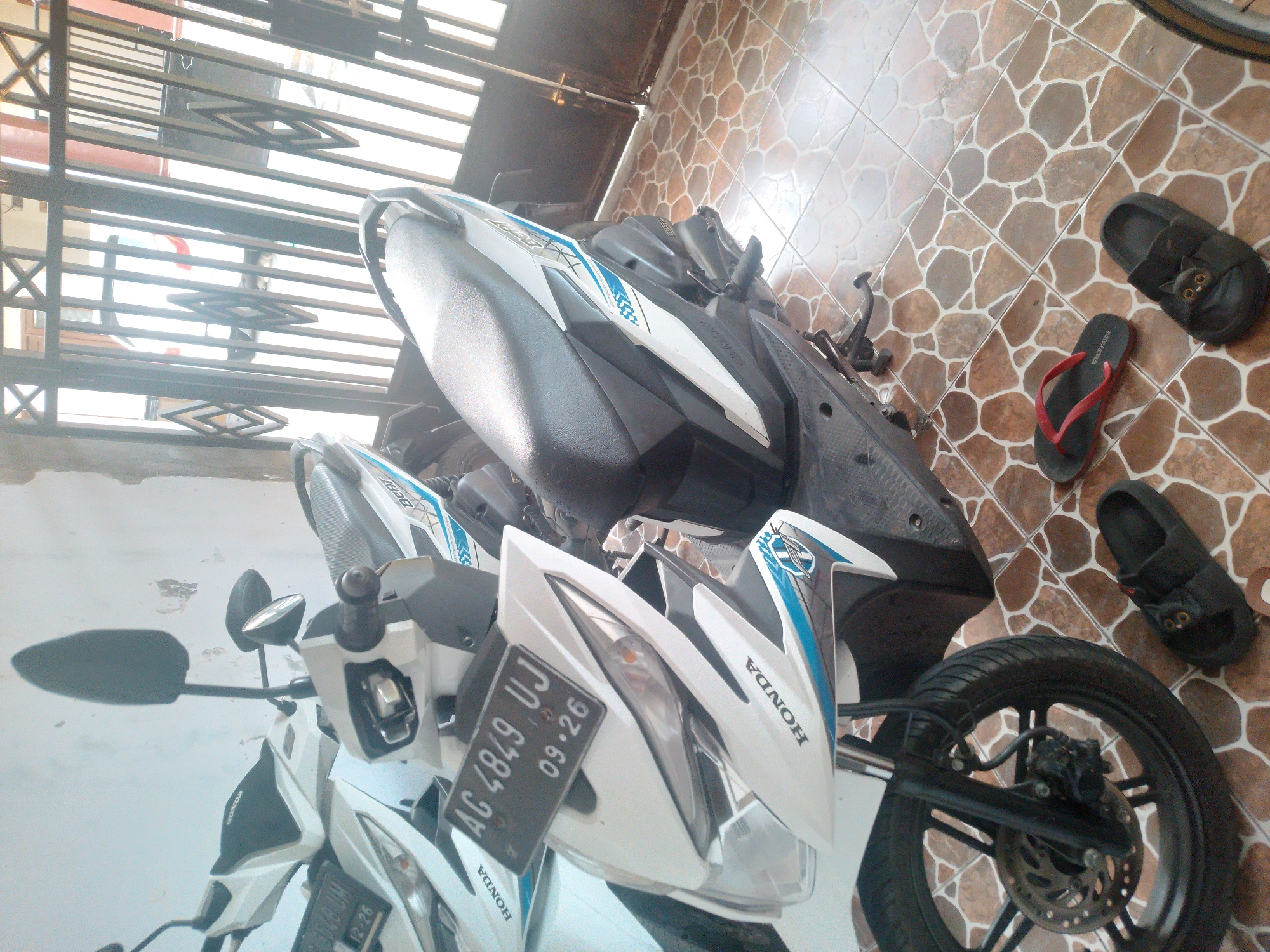 In Pelem, Indonesia, two white Honda motorcycles are parked on a tiled floor in front of a metal gate. The motorcycle in the foreground is closer to the viewer and has a license plate that reads "AG 4849 UJ 09.26". Both motorcycles have "HONDA" branding. There are three pairs of sandals on the floor: a pair of red flip-flops, a pair of black sandals with cat-face embellishments, and another pair of black sandals. The gate is open, revealing sunlight and a blurry view of a vehicle beyond. The environment suggests an outdoor area, possibly a garage or driveway.