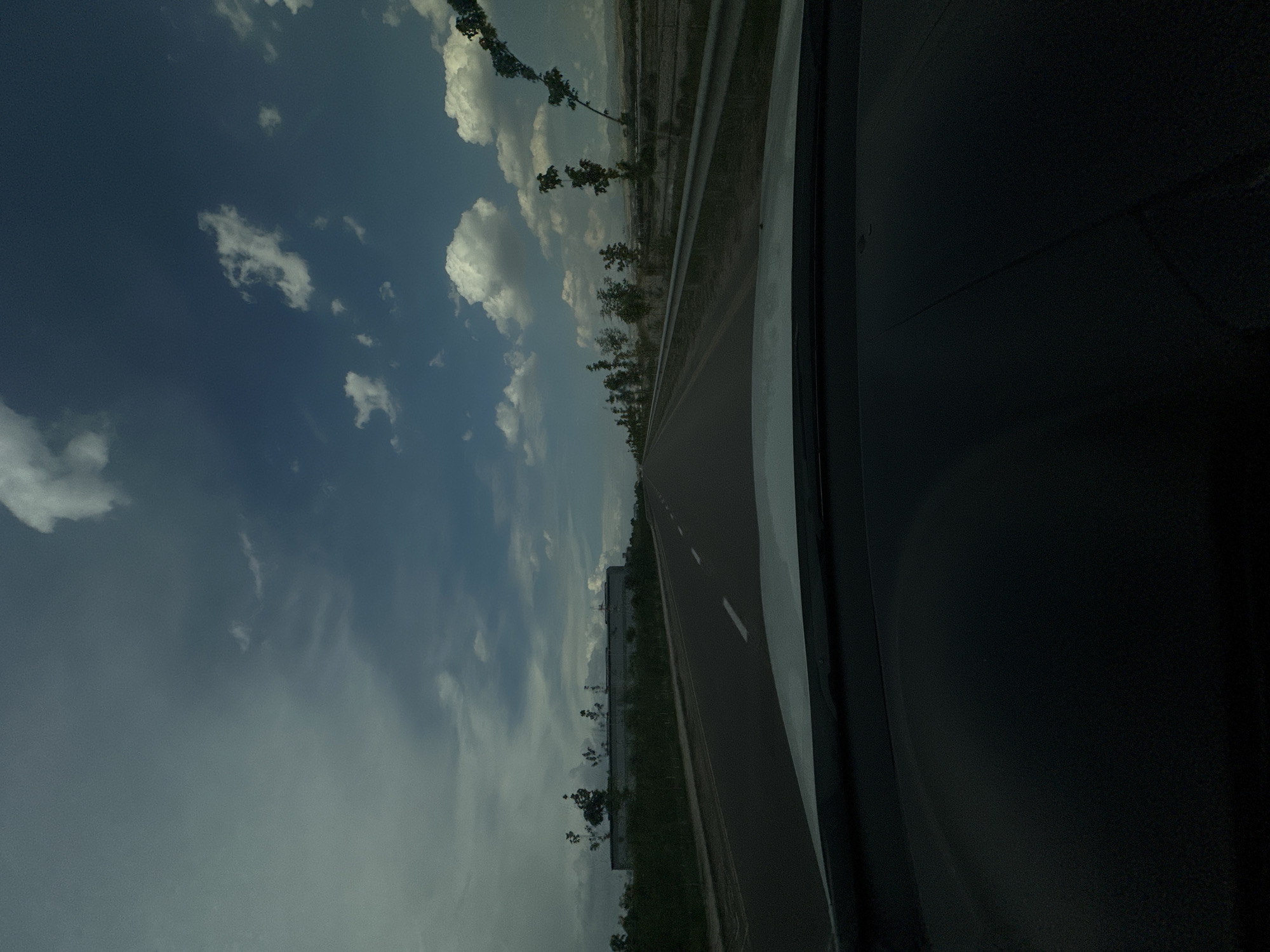 The image shows a view from inside a car looking out at a highway in Nalazar, Uzbekistan. The sky is cloudy with patches of blue sky visible. On the left side of the road, there is a long, low building with a fence in front of it, surrounded by green vegetation and several trees. On the right side of the road, there is a row of young trees lining the highway, with a metal guardrail separating the road from the adjacent landscape. The road itself is paved with white lane markings. The foreground is dominated by the dark interior of the car, including the dashboard and windshield. The overall scene depicts a typical highway scene in a semi-urban or rural area.