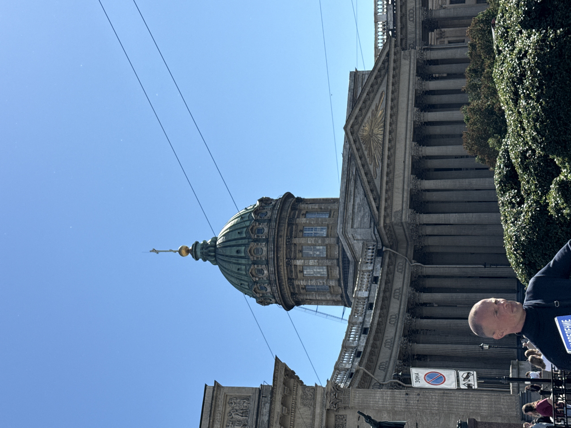 The image shows the Kazan Cathedral in Saint Petersburg, Russia, on a clear, sunny day. The dominant subjects are the grand architecture of the cathedral, including its iconic dome and colonnades, and several people visible in the foreground and background. A man in a dark jacket is looking towards the cathedral, with a blue sign visible on his person. Other people are also visible, some seated and others standing, suggesting a public gathering or a tourist area.
The scene is set outdoors, with the bright blue sky taking up a significant portion of the frame, indicating daytime. Overhead, electrical wires are visible, strung across the sky. Lush green foliage, possibly trees or ivy, covers a section of the building on the right side, providing a contrast to the stone architecture.
Notable details include the intricate details of the cathedral's facade, the classical columns, and the golden finial atop the dome. The presence of signage, including a traffic sign with Cyrillic text, confirms the location as Russia. The overall mood seems calm and observational, with the bright sunlight creating a pleasant atmosphere. The visible text on the blue sign worn by the man in the foreground appears to be in Cyrillic, though it is partially obscured.