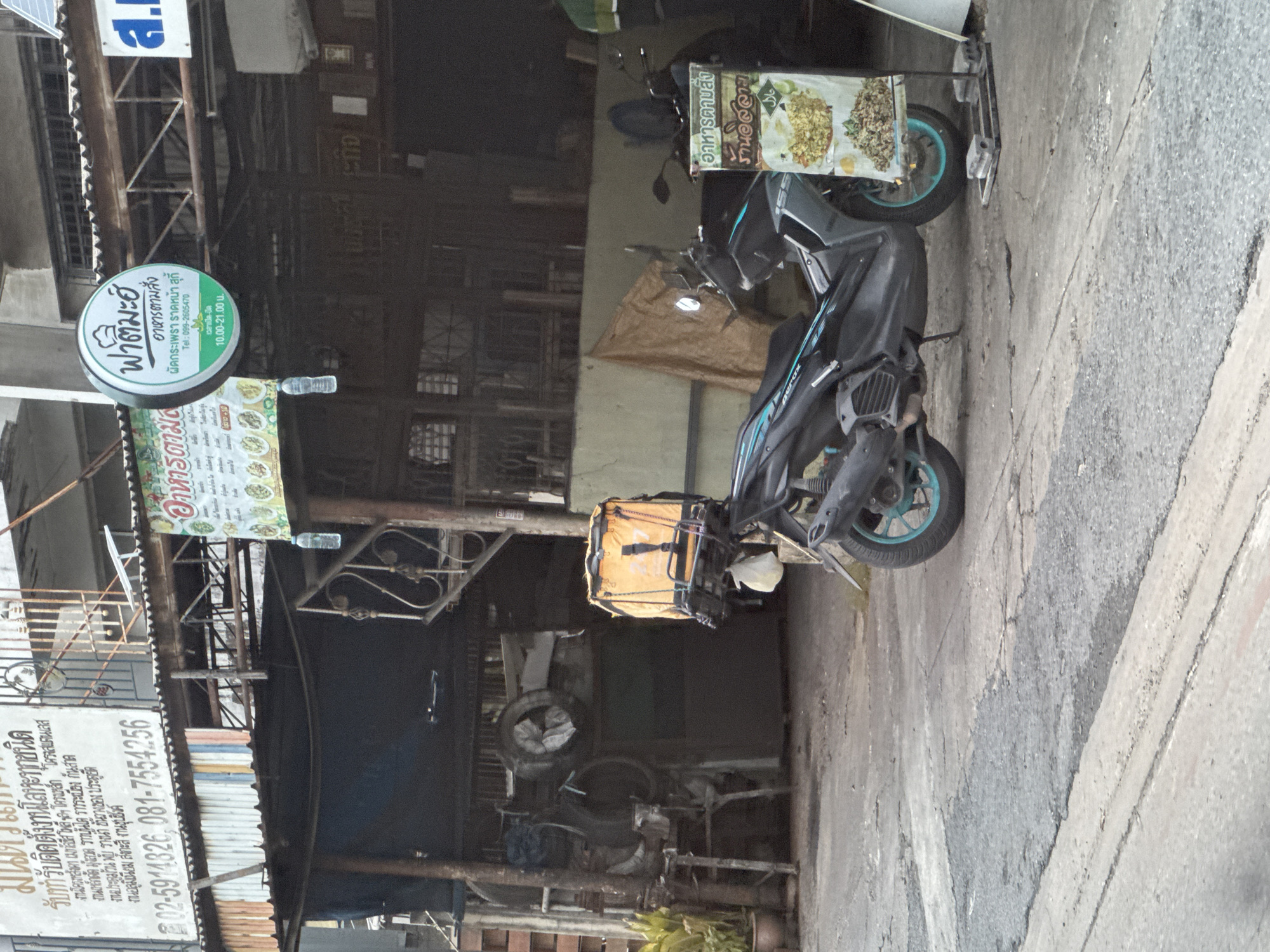 The image shows a street scene in the city of Nonthaburi City Municipality, Thailand. In the foreground, a black motorcycle with a teal-colored rim is parked on a cracked asphalt surface. Attached to the rear of the motorcycle is a bright orange delivery box. To the right of the motorcycle, a white stand displays a food menu with pictures of dishes and Thai text.
Behind the motorcycle, a covered outdoor area with a corrugated metal roof is visible. A large orange delivery bag is suspended from a rack in this area. Further back, there are structures with wooden supports and corrugated metal siding. Potted plants are present near the entrance to one of the structures. Various signs are displayed, including a circular green and white sign with Thai text and a smaller rectangular sign with a food menu. The overall environment suggests a commercial area, possibly a restaurant or food stall.