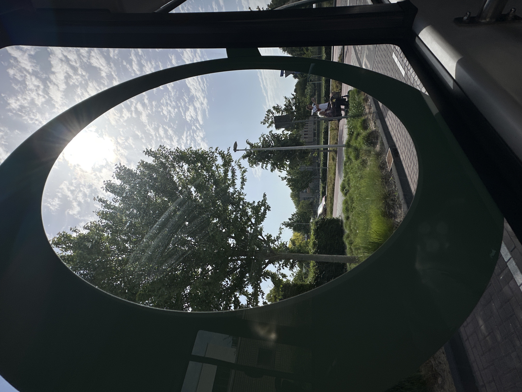 The image, taken from inside what appears to be a vehicle (possibly a bus or tram), shows a view of the outside world framed by an elliptical, green structural element. The scene depicts a typical Dutch urban landscape on a sunny day with scattered clouds. A large, leafy tree dominates the left side of the frame. A street and sidewalk are visible to the right of the tree, with pedestrians walking along it. A lamppost is also visible. The location is Dronten, Netherlands.