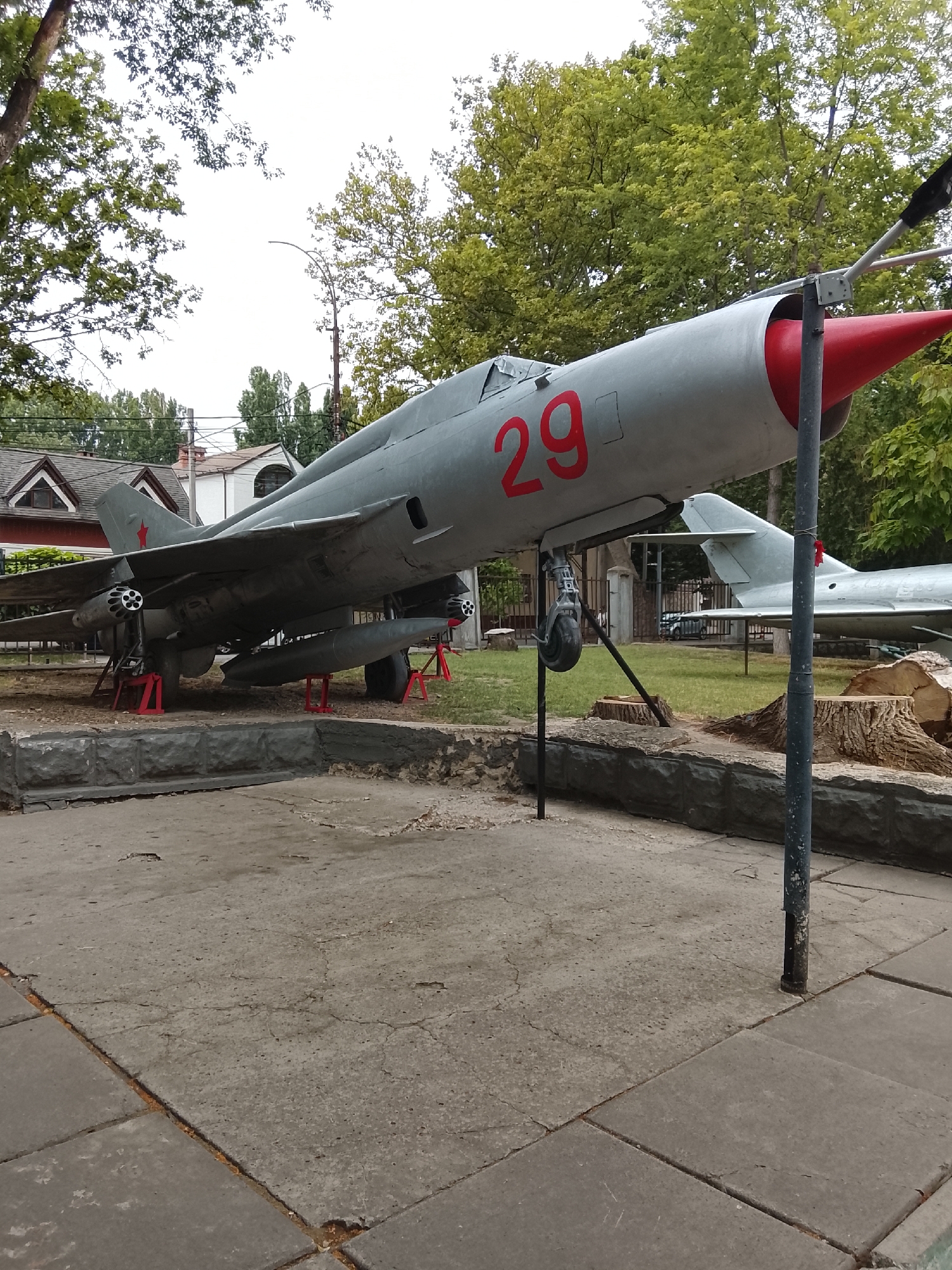 The image shows a parked MiG-21 fighter jet on display, likely at an open-air museum or memorial site. The aircraft is painted in a light gray camouflage, with the number "29" prominently displayed in red on its fuselage. A red cone-shaped nose further accentuates the aircraft's design. The jet is supported by red stands, indicating it is not operational. In the background, lush green trees and some buildings suggest an outdoor urban or suburban setting. The foreground consists of a concrete walkway and a low stone wall. There are no people visible in the image. The aircraft's military markings, specifically the red star on its tail fin, suggest a Soviet-era origin. While the exact location is not specified, the presence of a Soviet-era aircraft on display could be consistent with a military museum or a memorial in a city with a historical connection to the Soviet Union, such as Chișinău, Moldova.