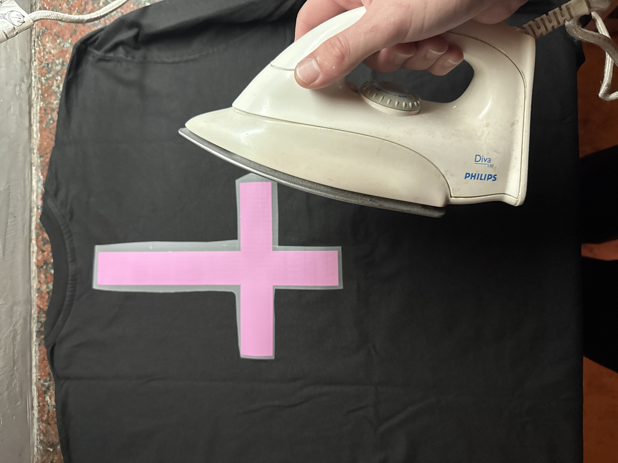 A hand holds a Philips Diva 130 iron over a black t-shirt with a pink cross design being ironed. The scene takes place indoors, possibly on a table or counter, but the location details of Buenos Aires, Argentina, are not discernible from the image.