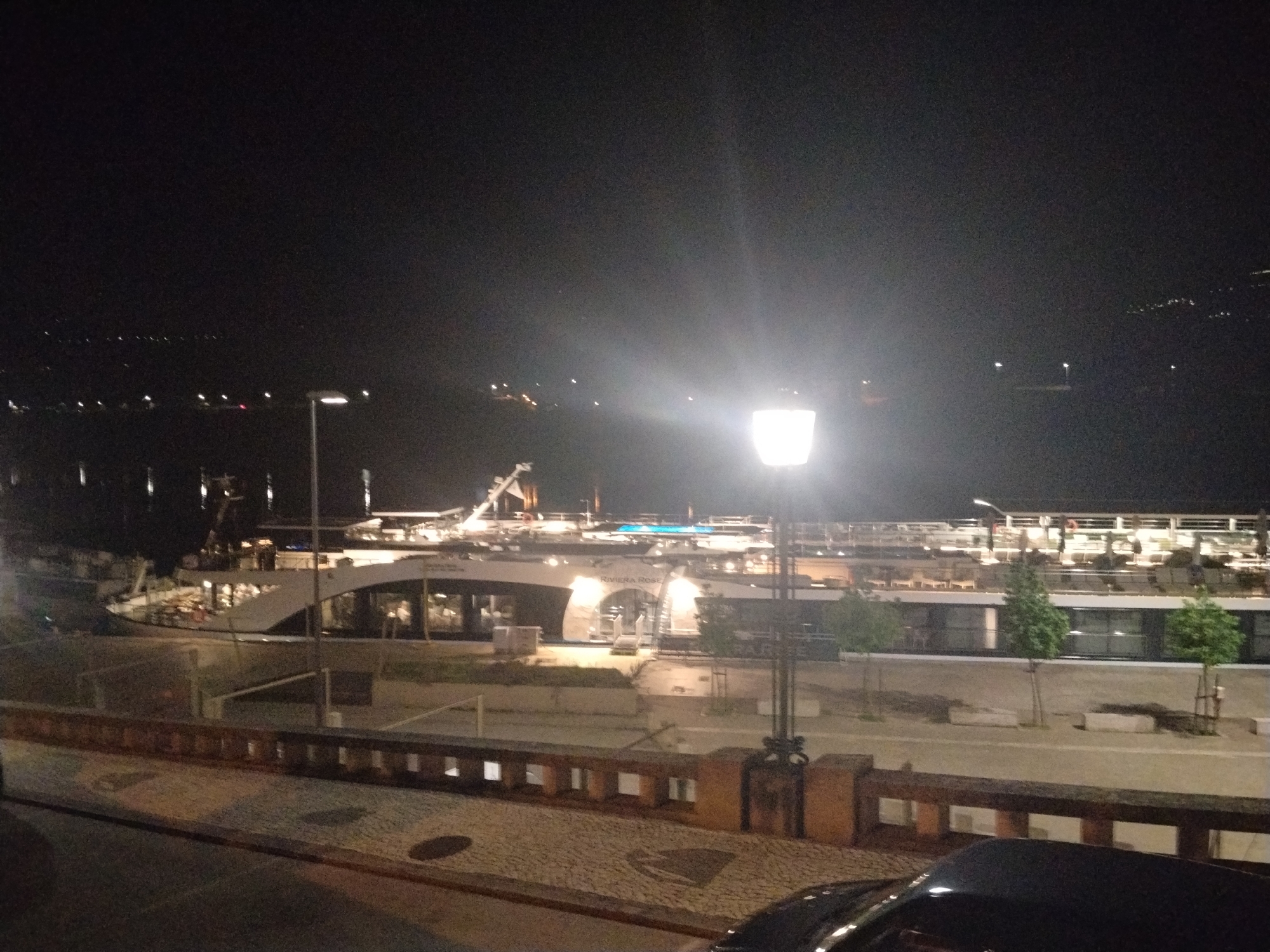 The image depicts a nighttime scene in Peso da Régua, Portugal. A riverboat, "Riviera Rose," is docked on the Douro River. A street lamp illuminates the foreground, casting light on a stone walkway with a low wall. Several small trees line the walkway. A dark-colored vehicle is partially visible in the lower right corner. Lights from buildings and distant hills are visible across the river. The sky is dark and clear.