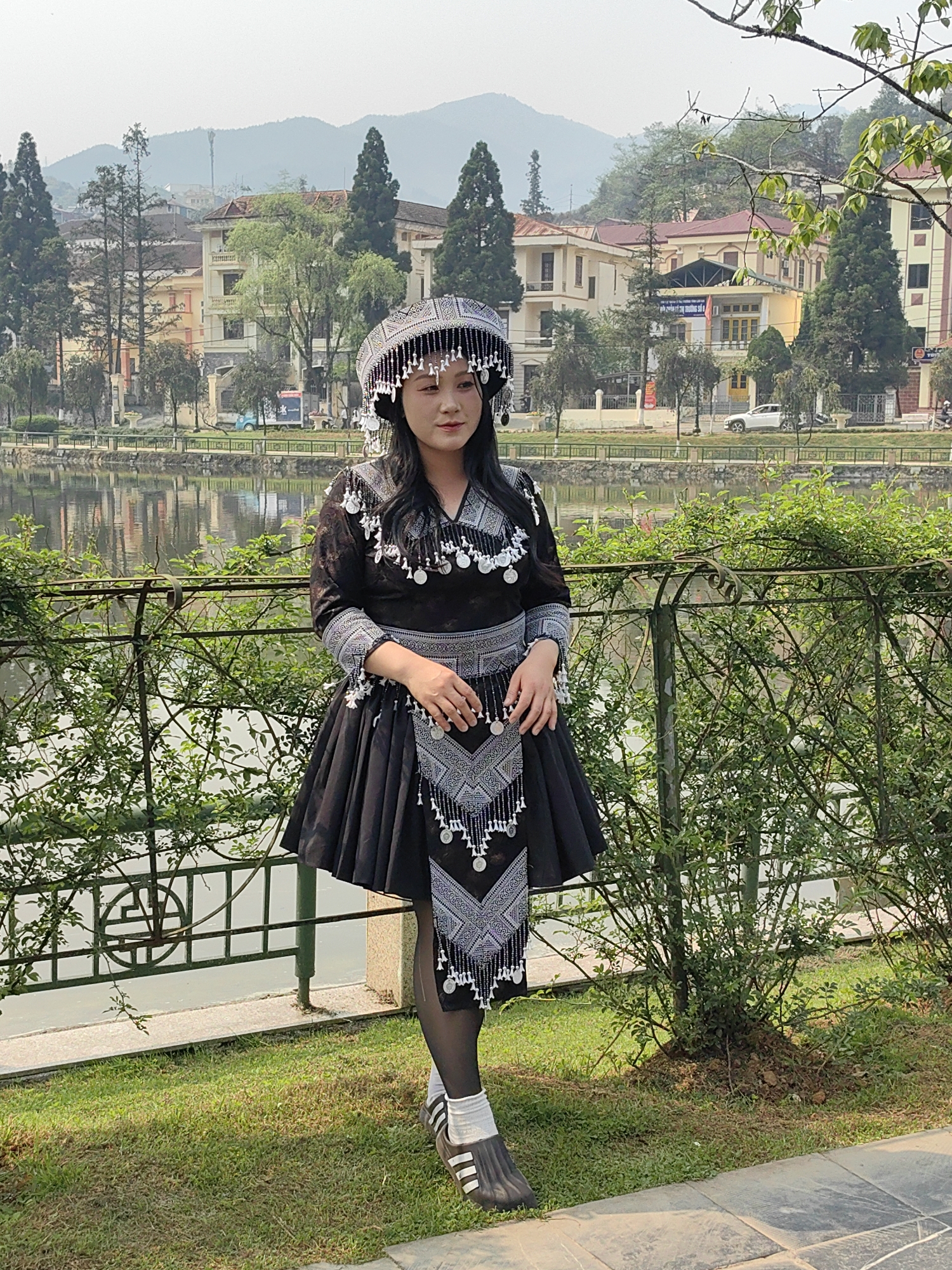 A young woman, likely of Hmong ethnicity, stands on a manicured grass lawn adjacent to a paved path, posing for a photograph. She is positioned outdoors near Sa Pa Lake in Sa Pa, Vietnam. The woman wears traditional attire consisting of a black long-sleeved top and a pleated skirt, both adorned with intricate silver geometric patterns and silver coin-like embellishments. A decorative silver-patterned panel with hanging tassels extends from her waist. She also wears a matching elaborate headdress with silver ornamentation and white tassels framing her face. Her footwear includes black tights, white socks, and dark slip-on shoes with white stripes.
Behind her, a dark green metal fence is partially obscured by dense green bushes. Beyond the fence, the calm surface of Sa Pa Lake reflects the sky and distant structures. Across the lake, a paved road is visible with several parked vehicles, including a white SUV and a light blue utility truck. In the background, multiple multi-story buildings, characterized by light-colored facades and varied rooflines, are nestled among numerous evergreen and deciduous trees. Prominent among these is a building with a yellow facade and a red roof. Distant mountains rise hazily under a pale sky, completing the urban-natural landscape of Sa Pa.