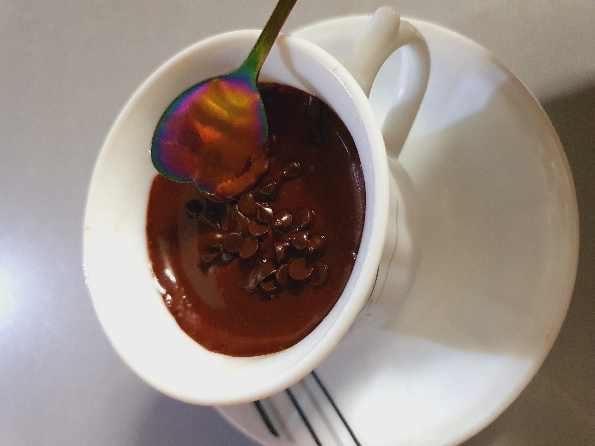 A white cup filled with dark chocolate pudding and topped with chocolate chips rests on a white saucer. A heart-shaped spoon with an iridescent finish is partially submerged in the pudding. The scene is a close-up shot, suggesting an indoor setting. There is no indication of the location being Shiraz, Iran, or any specific city.