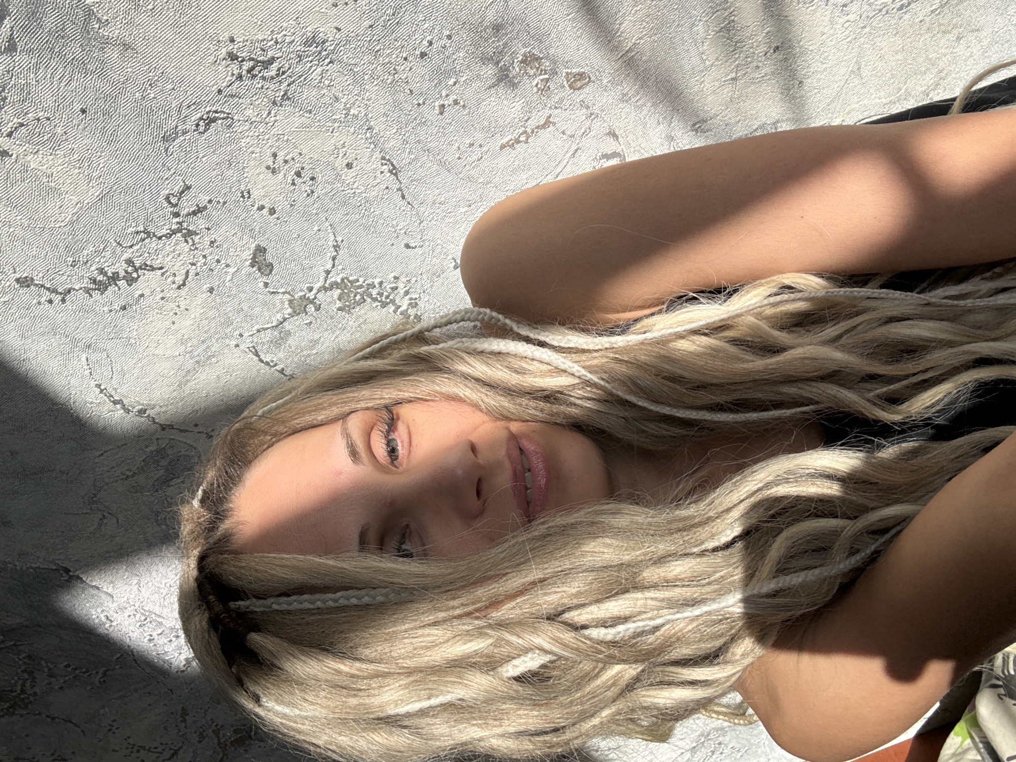 A woman with blonde, wavy hair mixed with white braids is lying down. Part of her face is illuminated by direct sunlight, while the other part is in shadow. She has fair skin, light eyes, and pink lips. She is wearing a dark-colored top. The background is a textured, light gray wall, with patterns resembling marbling. A shadow is cast on the wall. The image's location in Rusanovo, Russia, cannot be determined.