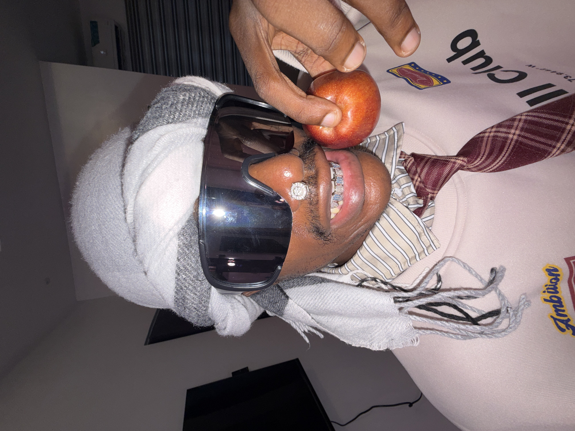 A man wearing sunglasses, a gray and white scarf, and a diamond ring on his nose holds an apple up to his face. He has a diamond-encrusted grill on his teeth and is wearing a tie and a light pink sweatshirt with the word "Ambition" on it. The photo was taken in Igboefon, Nigeria.
