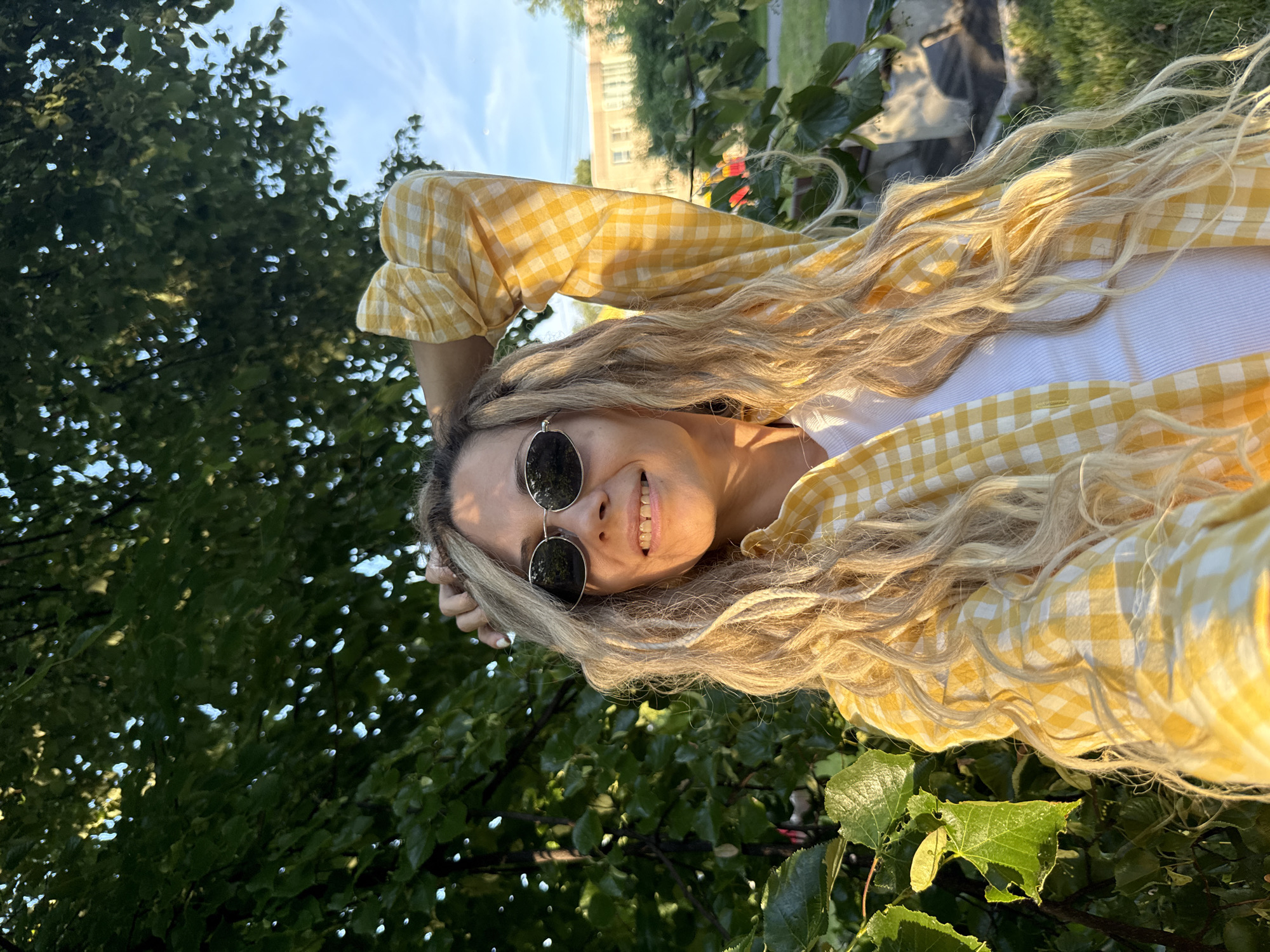 A blonde woman with long, wavy hair and sunglasses smiles as she holds her arm over her head, posing for a selfie in front of a lush green tree. She is wearing a yellow and white checkered shirt over a white tank top. The sun is setting, casting a warm glow on her hair and face. The background shows a blue sky with scattered clouds and a building in the distance. The scene is set outdoors in a park or backyard.
