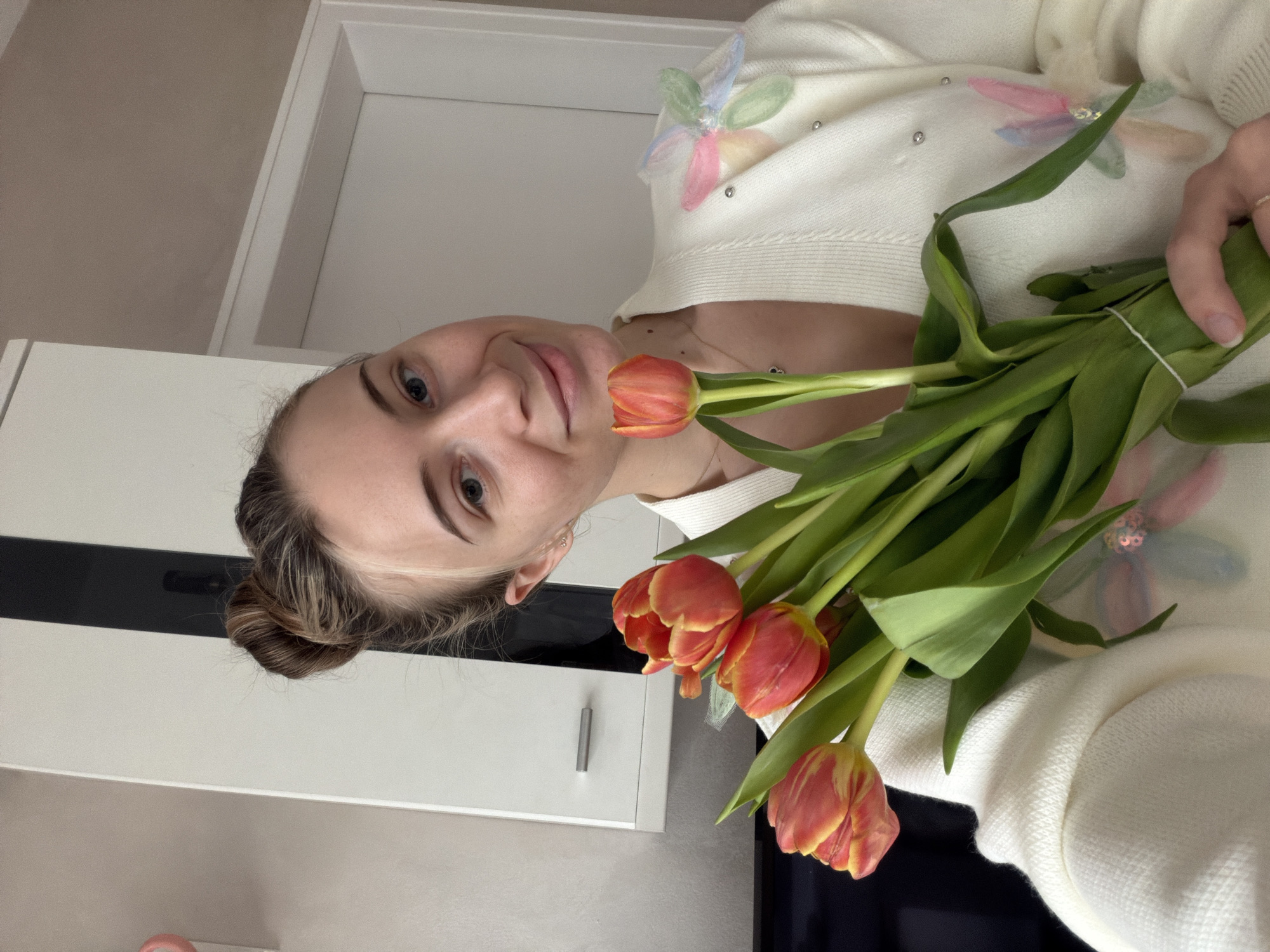 A woman holds a bouquet of three red-orange tulips. She wears a white sweater decorated with pastel-colored, flower-like embellishments. Her hair is styled in a bun. The background includes white kitchen cabinets with black accents. The location is not discernible from the image.