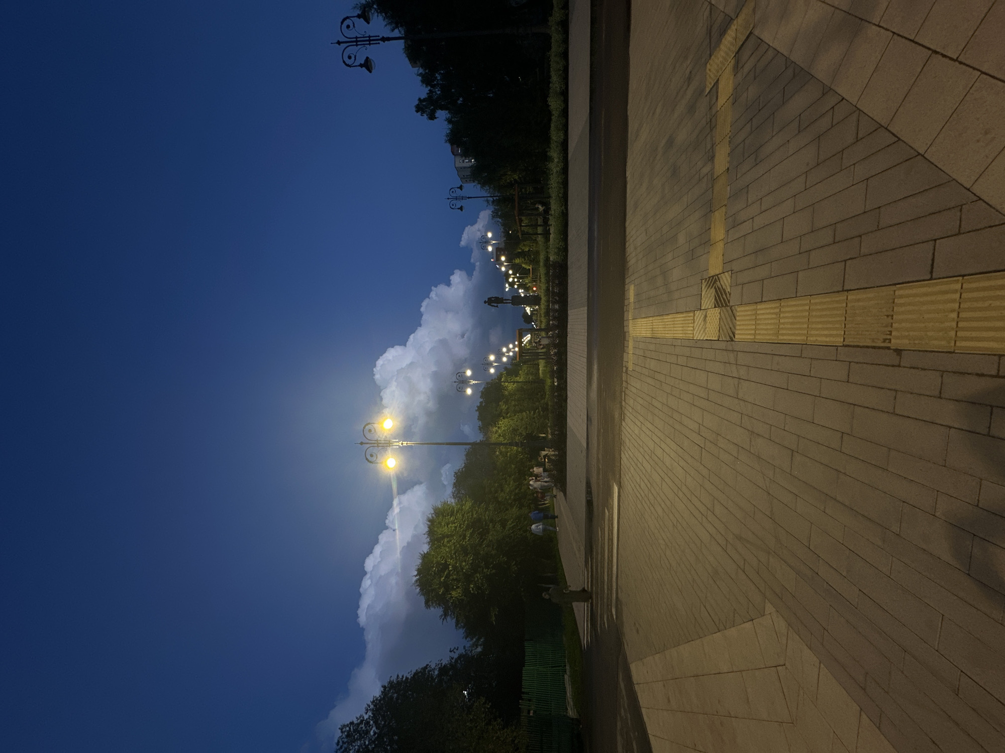 The image depicts an exterior scene at dusk in Kokshetau, Kazakhstan. The foreground is dominated by a paved walkway with a yellow tactile paving strip. In the midground, a road and a crosswalk are visible, with a pedestrian crossing. Beyond the crosswalk, a park area is illuminated by streetlights, revealing lush green trees, some with distinct fluffy white clouds hanging in the deep blue sky above them. Several people are walking in the park. In the distance, a statue of a person stands prominently, and apartment buildings are visible through the trees. The overall atmosphere is calm and serene, with the artificial lights casting a warm glow on the surroundings.