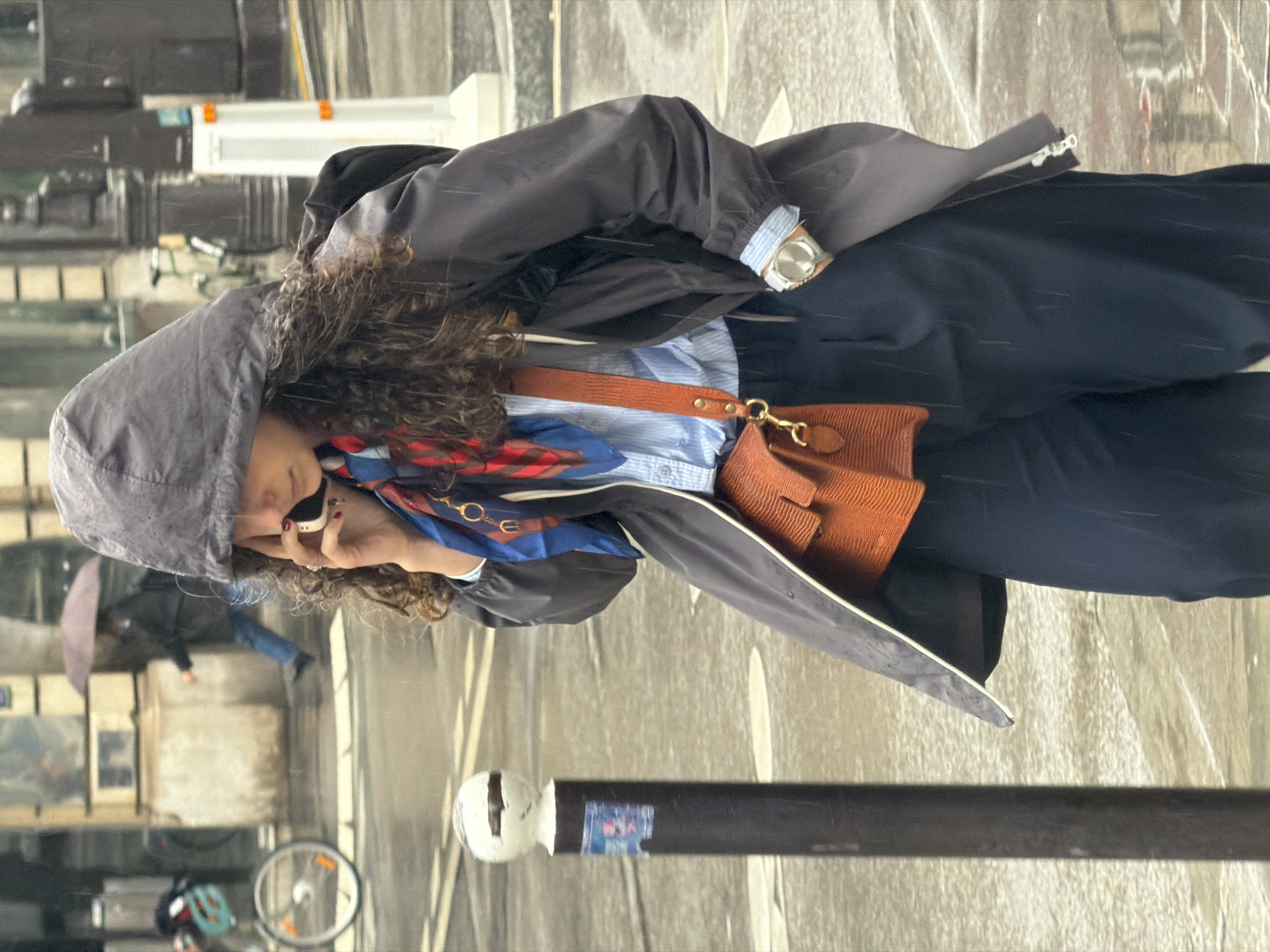 A woman with curly brown hair stands in the rain on a Parisian street, holding a cell phone to her left ear. She is wearing a gray hooded raincoat, a blue and red patterned scarf, a light blue striped shirt, navy slacks, and a brown shoulder bag. A wristwatch is visible on her left wrist. The street is wet and reflective, suggesting it has been raining for a while. In the background, a person carrying an umbrella walks by, and a bicycle is parked. A metal bollard stands in the foreground. The setting is urban, likely a commercial street in Paris, France.