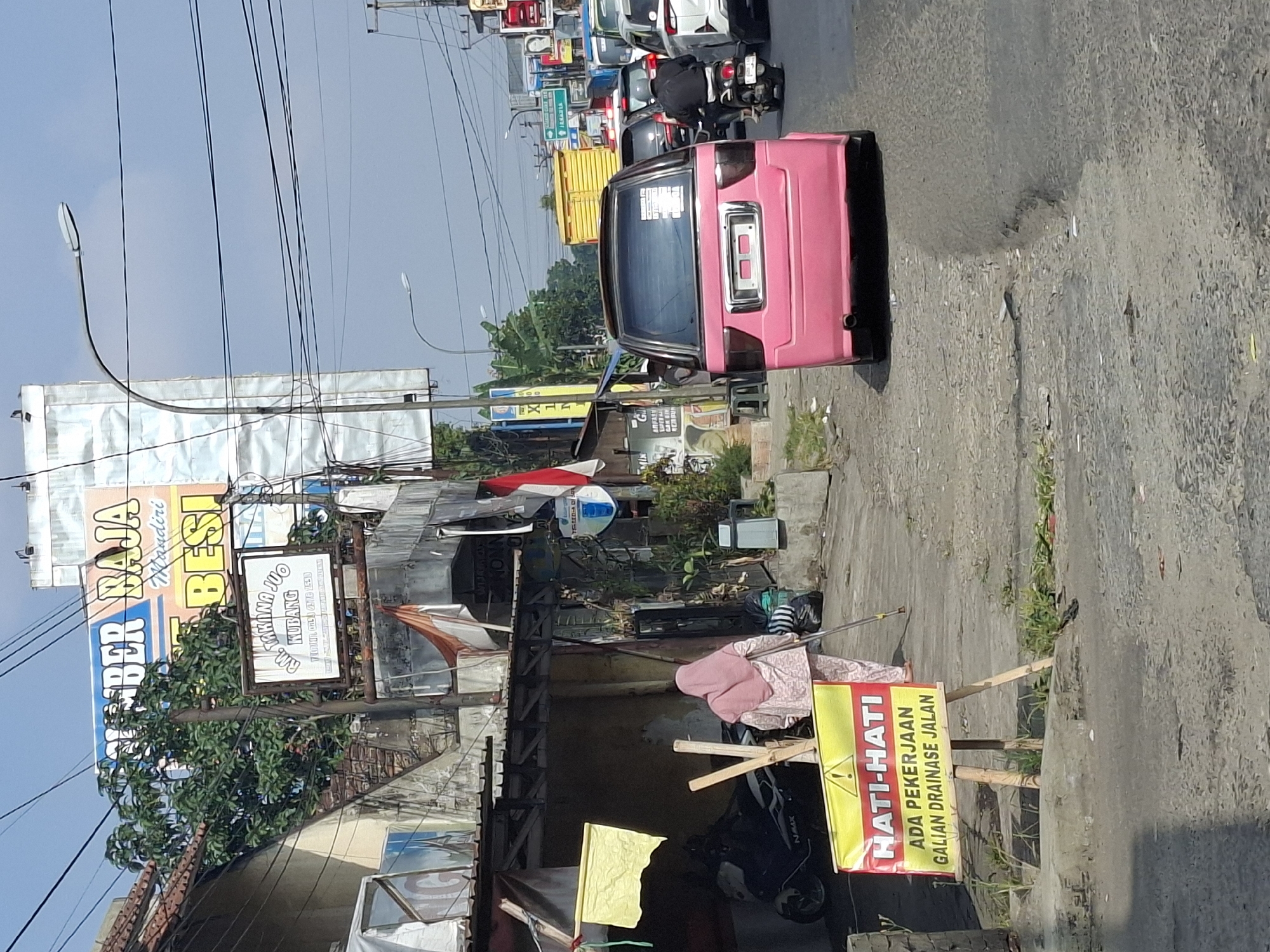 The image captures a street scene in Sukaraja, Indonesia, on a sunny day. A pink van is the central subject, parked on the right side of the road, facing away from the camera. To the left of the van, a person wearing a pink hijab and a matching patterned outfit is walking away from the viewer, leaning on a walking stick. In front of the person is a yellow sign with red lettering that reads "HATI-HATI ADA PEKERJAAN GALIAN DRAINASE JALAN," which translates to "BE CAREFUL THERE IS ROAD DRAINAGE EXCAVATION WORK."
The environment shows a mix of urban elements. Buildings line the left side of the street, with signage visible, including a large blue and orange sign that says "SUMBER BAJA MANDIRI BESI". A street lamp is positioned on the left, and overhead wires crisscross the sky. The road surface appears to be asphalt, with some visible wear and tear. Traffic is present, with other vehicles, including cars and a motorcycle, visible in the background, suggesting moderate congestion. Green trees and vegetation are interspersed among the buildings and along the roadside. The overall impression is of a typical urban street in a developing area, with ongoing infrastructure work causing a potential hazard.