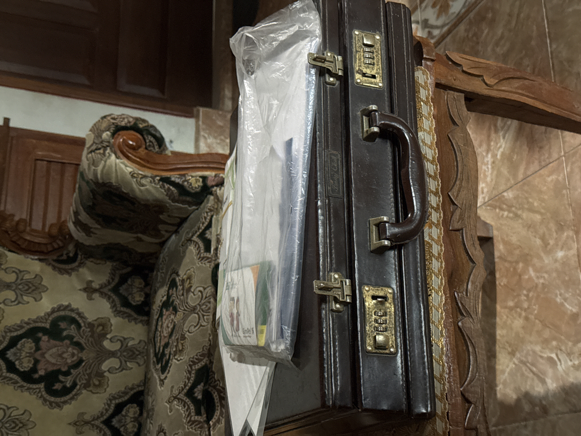 In the city of Bireuenmeunasahblang, Indonesia, a dark brown leather briefcase with brass accents and a combination lock sits on a carved wooden table. The briefcase is topped with a clear plastic-wrapped stack of papers. Behind the briefcase, a patterned sofa with a wooden armrest is visible. The floor appears to be tiled.