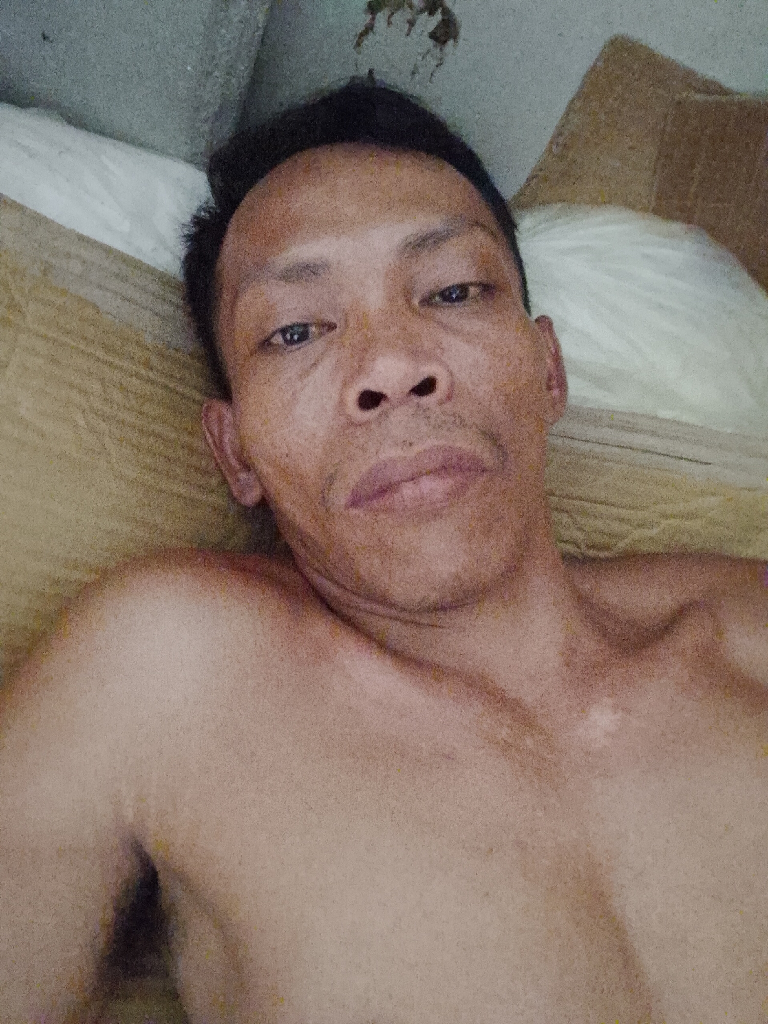 A shirtless middle-aged man with short black hair is lying down indoors. His skin tone appears to be brown. He is resting against a light brown surface, potentially cardboard, with a white pillow partially visible behind his head. The background includes a gray wall or ceiling. The image appears to have been taken from a close-up, overhead perspective. Location: Unspecified location in Bukit Ketok, Indonesia.
