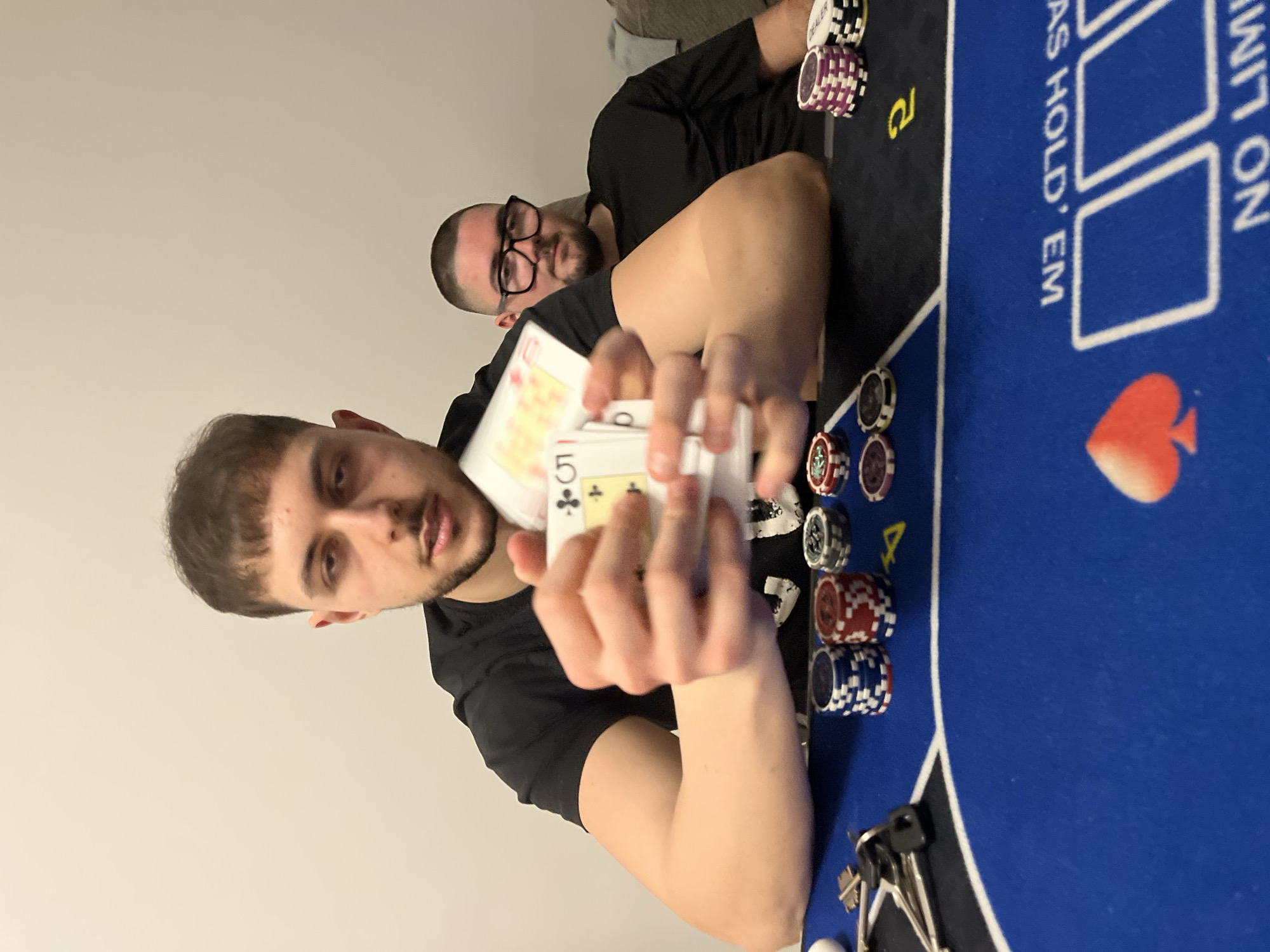 An image, rotated 90 degrees counter-clockwise, captures two men playing poker indoors in Burgas, Bulgaria. The shot is taken from a slightly elevated perspective, looking down at the table.
In the foreground, a young man with short dark hair and a light beard, wearing a black t-shirt, holds a hand of playing cards. His hands are visible, and the cards show a '10' of hearts and a '5' of clubs, along with other partially obscured cards. He looks towards the camera with a neutral expression.
Behind him, to the right of the frame, another man with short hair or bald, glasses, and a beard, also in a black t-shirt, is visible. He is looking towards the foreground man, seemingly observing the game.
The central object of the scene is a blue poker table with black borders and white markings. Several stacks of poker chips in various colors—white, purple, black, red, and blue—are arranged on the table. A white "DEALER" chip is visible near the man in the background. Printed text on the blue felt reads "AS HOLD 'EM" and partially "NO LIMIT ON," indicating a "No-Limit Hold'em" game. A red spade symbol is also visible on the table. A set of keys rests on the table in the bottom right corner. The background consists of a plain, light-colored wall. The overall mood is casual and focused on the game. The time of day is not discernible but is likely indoors.