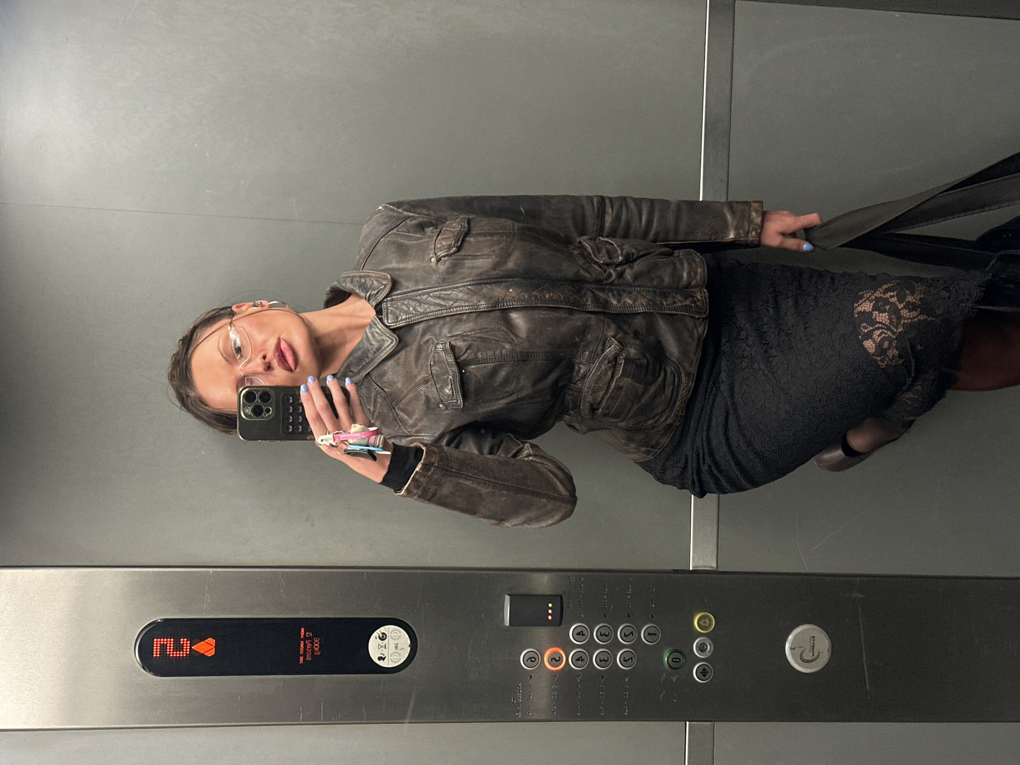 A woman is taking a selfie in an elevator. She is wearing glasses, a brown leather jacket, and a black lace dress. She is holding a black smartphone in her left hand, which has light blue nail polish on the nails. She has a key set attached to her phone. The elevator panel shows the current floor as "2" and the weight capacity as "800kg". The elevator interior is gray. There is no indication of this image being taken in the city of London, United Kingdom.