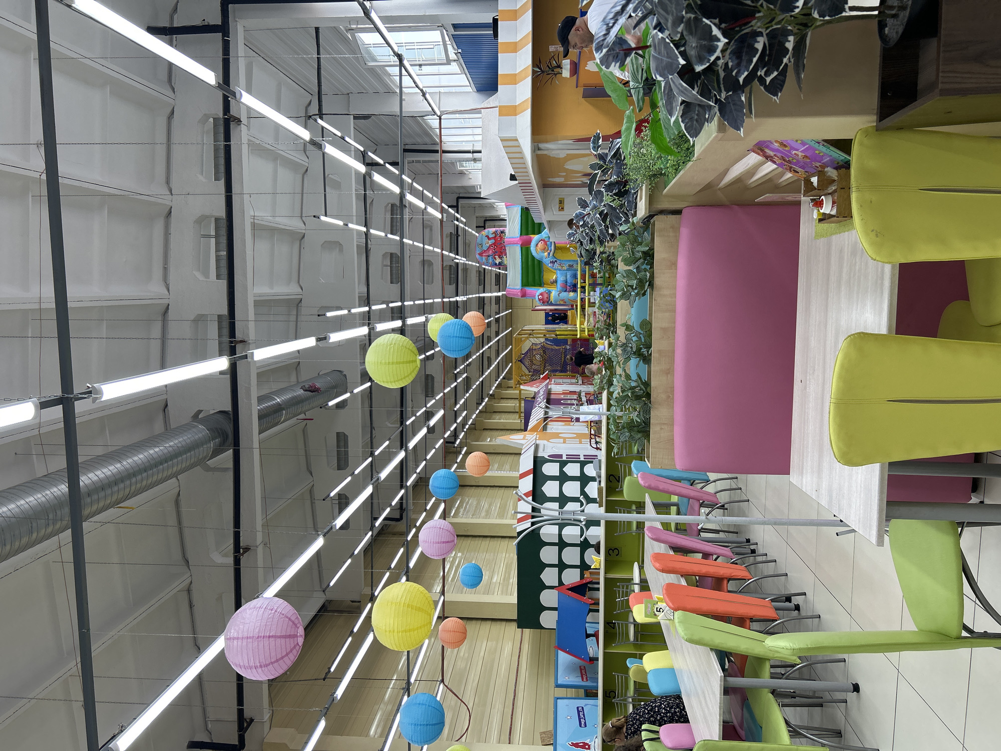 The image depicts the interior of a children's entertainment center in Chaikovsky, Russia. The scene features a bright, well-lit space with a high ceiling, adorned with numerous fluorescent light fixtures and industrial ventilation ducts. Hanging from the ceiling are colorful paper lanterns in various shades of pink, yellow, and blue, adding a festive atmosphere.
In the foreground and mid-ground, several tables and chairs are arranged, suggesting a dining or seating area. The chairs are a mix of vibrant colors, including lime green, bright orange, pink, and light blue. Some tables are set with what appear to be condiment caddies containing bottles and small containers. Behind these seating areas, a children's play area is visible, complete with colorful structures, inflatable slides, and various play equipment, some featuring cartoon characters.
Several individuals are present, though their interactions are not clearly discernible. In the left mid-ground, a woman is seated at a table, looking down. Further back, other people are visible within the play area, some appearing to be children and adults supervising them. In the right mid-ground, a man wearing a black baseball cap and a white t-shirt is partially visible, facing away from the camera.
The environment is characterized by a mixture of industrial elements (ceiling, ducts) and child-friendly aesthetics (bright colors, play structures, lanterns). The overall impression is that of a lively and engaging space designed for children's amusement and recreation.