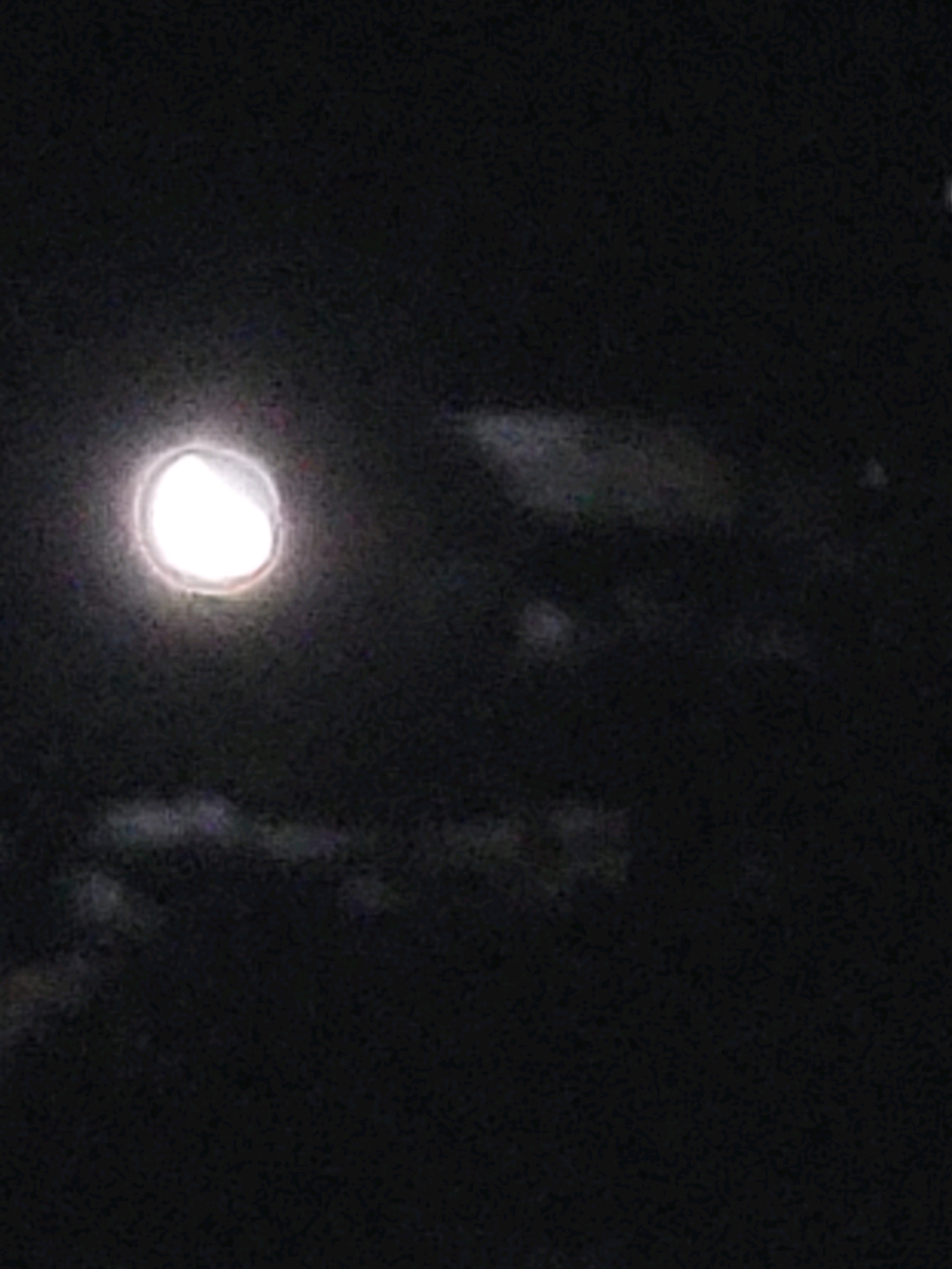 The image depicts a nighttime scene with a large, bright, circular object, likely the moon, dominating the left side of the frame. The object is partially obscured by clouds or atmospheric haze, giving it a diffuse glow. The background is dark, suggesting a rural or natural environment, possibly Ozolnieki, Latvia, during the night. There are faint, indistinct shapes in the lower portion of the image that could be trees or other landscape features, also rendered dark by the low light conditions. The overall impression is one of a clear night sky with a prominent celestial body.