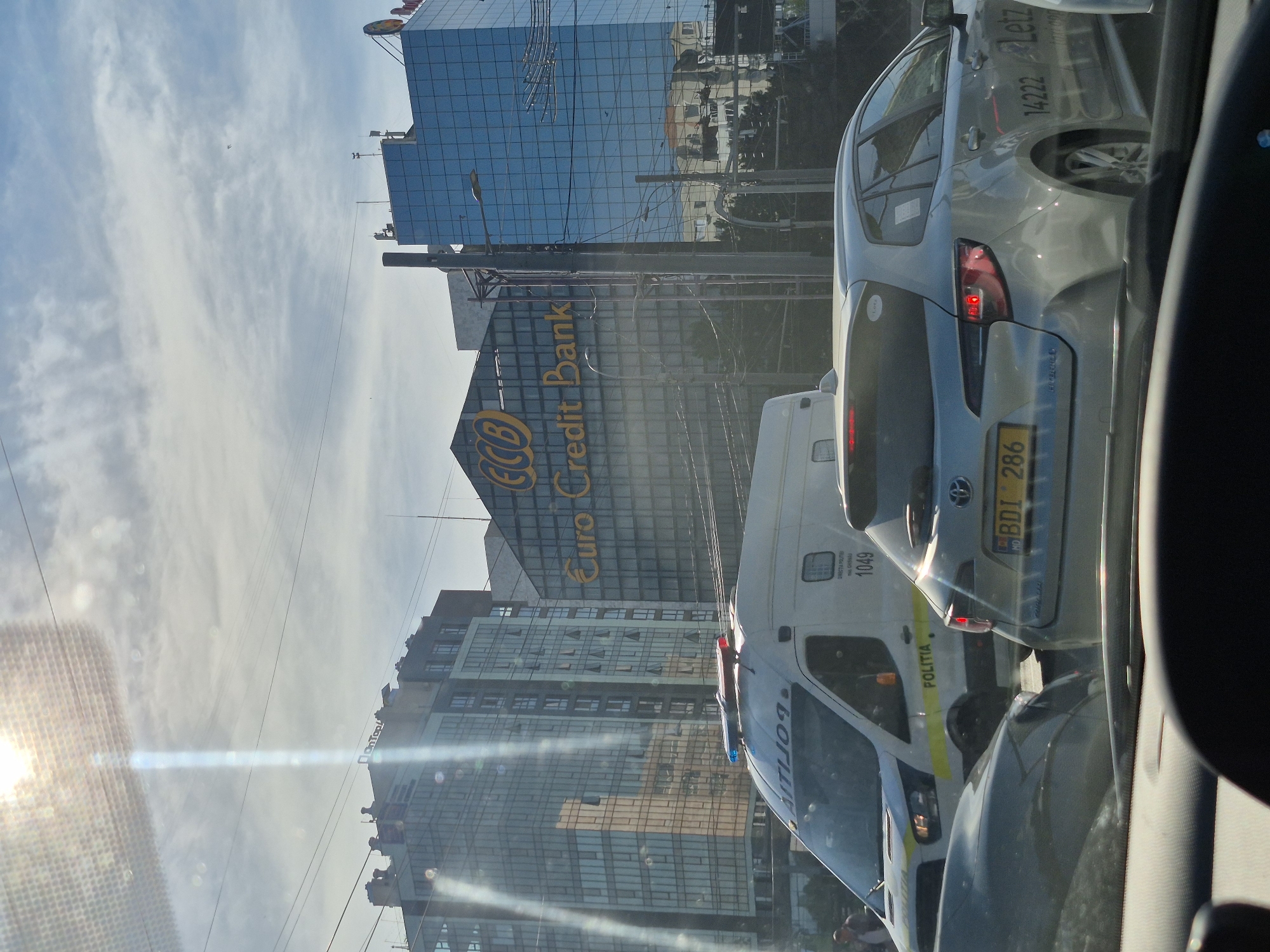 The image depicts a street scene in Chișinău, Moldova, on a partly cloudy day. In the foreground, the dashboard of a vehicle is visible, with reflections from the sun appearing on the windshield. Several cars are on the road, including a white police van with "POLITIA" written on its side and illuminated blue and red lights on its roof. A silver sedan with a license plate reading "BDI 286" is positioned behind the police van.
In the background, several modern buildings rise. One prominent building features a large sign for "ECB Euro Credit Bank." The architecture of the buildings suggests an urban commercial or financial district. Overhead, power lines and street lamps are present. The overall scene captures a moment of typical city traffic, with official vehicles and commercial buildings forming the backdrop.