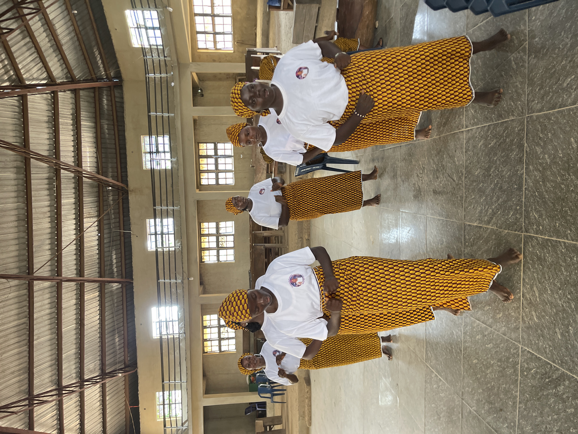 The image depicts an indoor scene within a large, unadorned hall in Ube, Nigeria, where at least seven adult women are engaged in a synchronized activity, possibly a dance or performance. All women are uniformly dressed in white short-sleeved t-shirts featuring a circular emblem on the chest, paired with long, patterned skirts or wraps and matching head coverings made from a distinct yellow fabric with a repeating brown oval design. They are all barefoot.
The women are positioned in rows, with some facing forward and others slightly angled, indicating movement. Their hands are generally clasped or positioned near their lower torsos. The woman in the foreground on the right side has a beaded bracelet on her left wrist.
The building features a high, exposed corrugated metal roof supported by reddish-brown metal trusses. The walls are light-colored, possibly beige or cream-colored, with a textured finish. Numerous rectangular windows with visible panes are set into the walls at different levels, providing natural light, some showing bright exterior light. An elevated balcony or mezzanine level with a dark metal railing extends along the upper part of the hall's perimeter. The floor is covered with large, square, grey-brown tiled flooring. In the background, scattered blue plastic chairs and wooden benches or furniture are visible, suggesting the space serves as a community or assembly hall. Bare light bulbs hang from the ceiling structure.