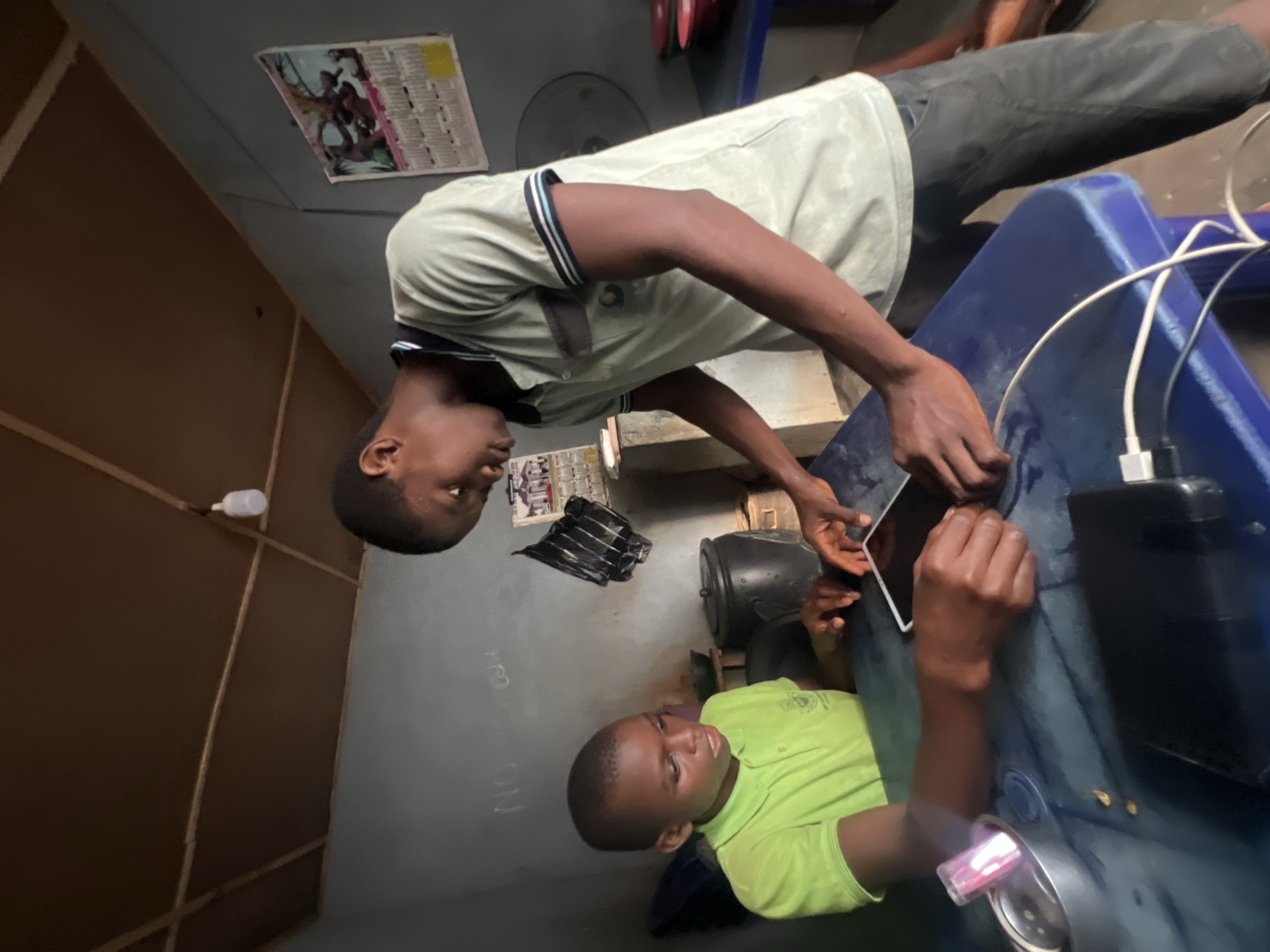 Two young Black males are engaged with a tablet on a blue table inside a dimly lit room. The male on the left, facing the viewer, wears a bright green polo shirt and looks intently at the tablet. The male on the right, standing and partially obscuring the left male, is dressed in a light grey polo shirt with dark trim and is also looking at the tablet, with his hands near it. Wires and a black power bank are on the table. The room has a brown, slatted ceiling with a light fixture. The walls are grey, with some markings and a calendar or poster visible. A large black pot and other objects are in the background. The setting appears to be a modest interior, possibly a home or small business, in Nkpor, Nigeria.