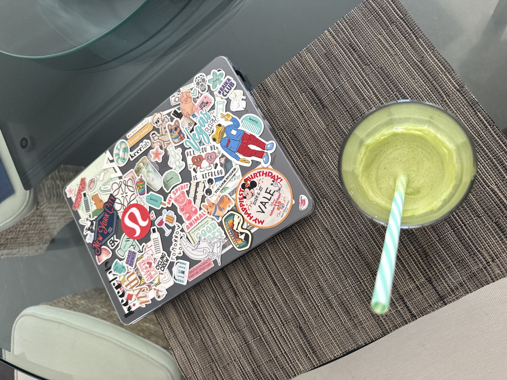 The image shows a laptop and a glass of green smoothie on a table. The laptop is covered in stickers, including brands and labels like "New York City", "Lululemon", "Keerlab", "Aerie", "Disney" and "Breckenridge Colorado." The green smoothie has a green and white striped straw in it. The table has a woven placemat. The image does not provide any information that can be used to identify the city of San Bernardino Tlaxcalancingo, Mexico.