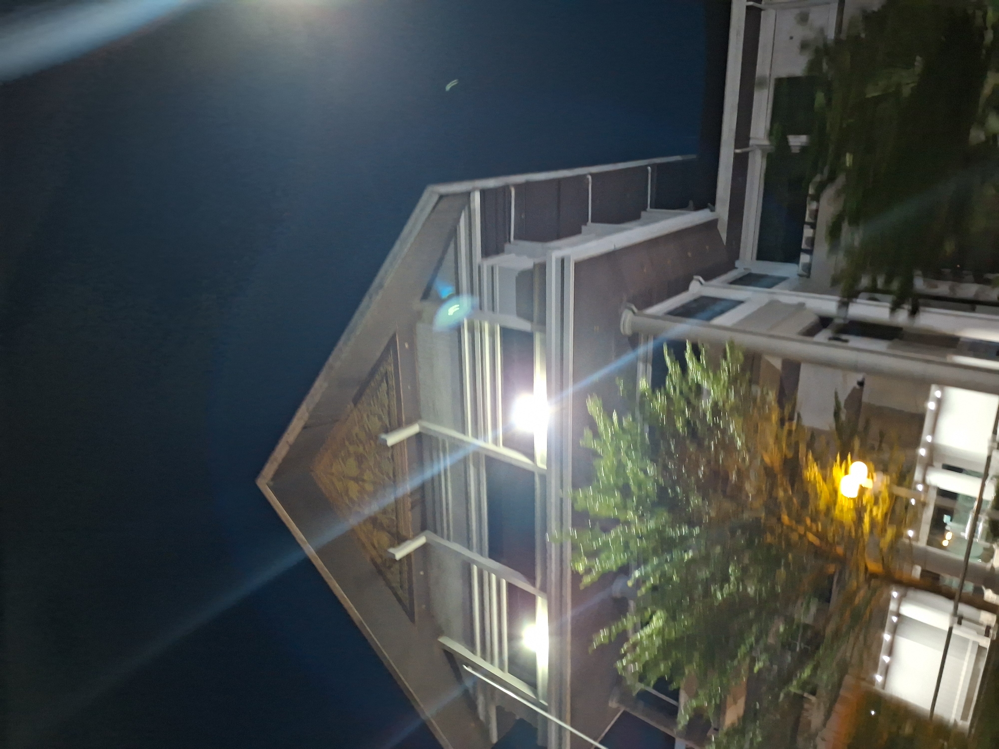 Here's a description of the image:
The image depicts a building at night in Lhokseumawe, Indonesia. The building has a prominent triangular facade with ornate decoration. The facade is partially illuminated by artificial lights that appear to be attached to the building. The building seems to be a commercial property. A large tree or bush partially obscures a portion of the building's front. The night sky is a dark blue, with some light glare.