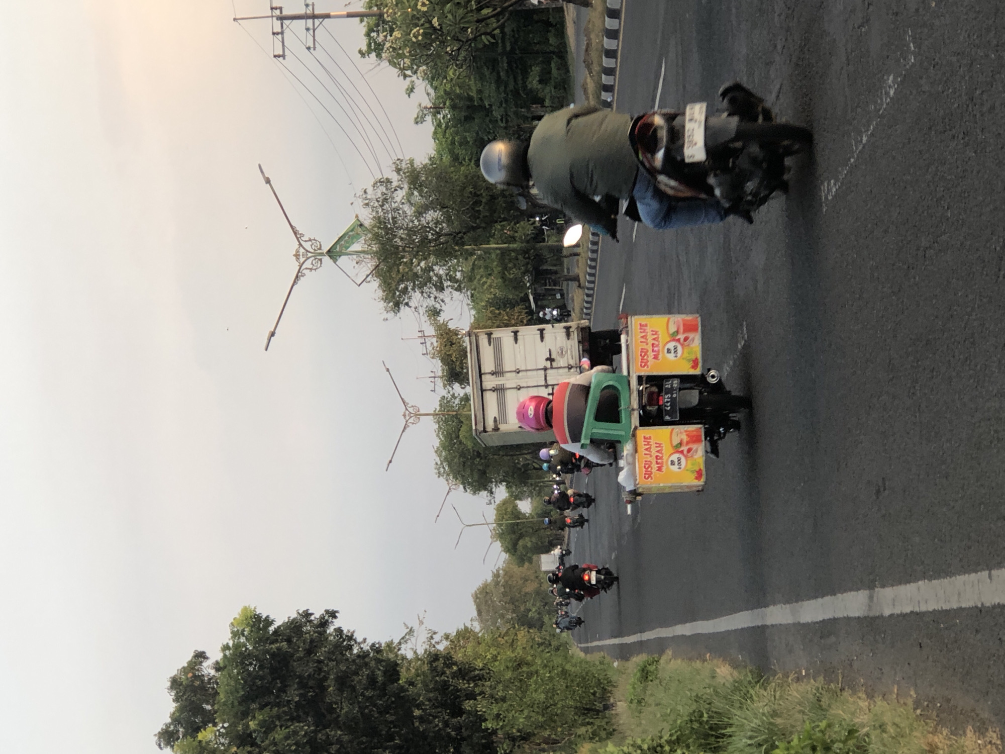 The image depicts a street scene in Siring, Indonesia, during daylight hours. A line of motorcycles is traveling on a paved road. In the foreground, a motorcycle carries a cart adorned with yellow signage that reads "SUSU JAHE MERAH" (Red Ginger Milk) and shows images of drinks. The rider of this motorcycle is wearing a pink helmet and a green vest. To the right, another motorcycle is visible from behind, with its rider wearing a gray helmet and a green jacket. A large white truck is positioned on the right side of the road. The road is lined with trees and vegetation on the left and right sides. Electrical poles and wires are visible in the background. The sky is overcast with a hint of sunlight towards the right.