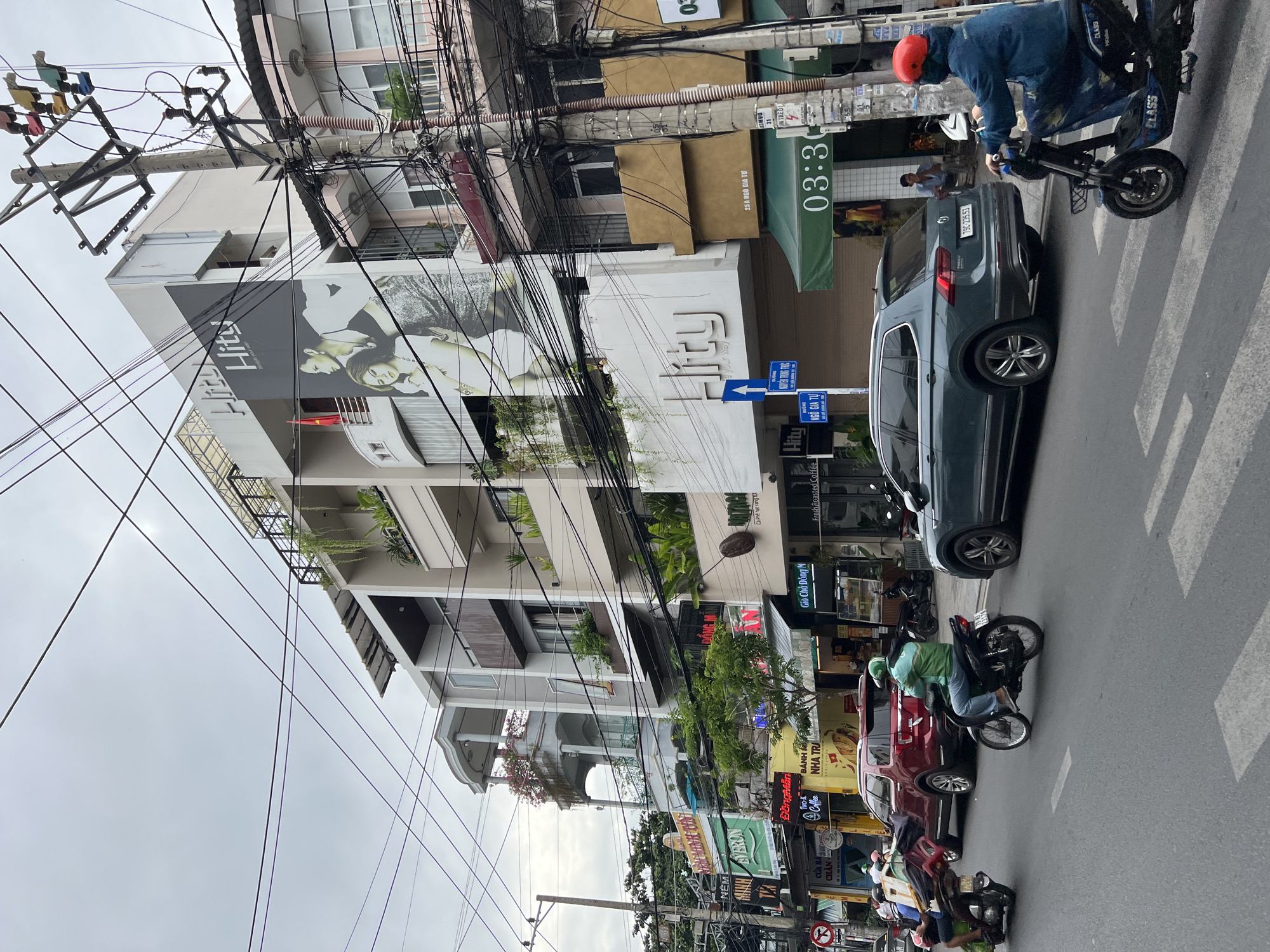 This image captures a street scene in Nha Trang, Vietnam, on an overcast day. The foreground shows a paved road with white pedestrian crosswalk markings. Several vehicles are present: a dark teal SUV in the right lane, a red SUV in the left lane, and multiple motorcycles with riders. One motorcyclist in the foreground wears a blue jacket and red helmet, riding an electric scooter across the crosswalk. Another motorcyclist in the left lane wears a green jacket and helmet.
The buildings lining the street are multi-story structures with modern architectural elements. The building on the right features a large mural of two people and the text "Hity lifestyle your way." Electrical wires are strung extensively across the scene, attached to utility poles. Signs are visible on the buildings, some in Vietnamese. One sign indicates "03:30." The environment appears urban, with a mix of commercial establishments and residential buildings. The sky is cloudy, indicating subdued lighting.