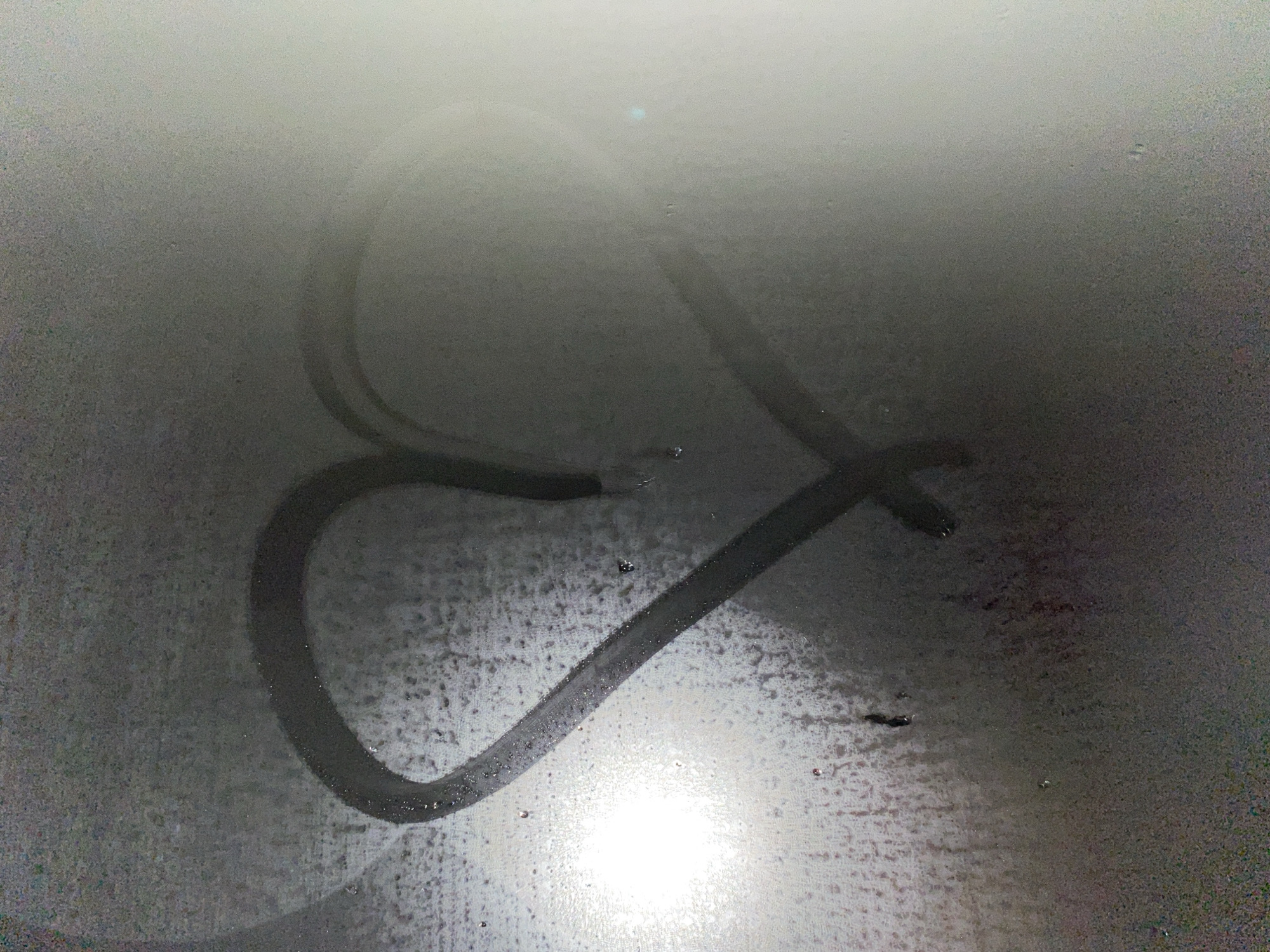 The image displays a heart shape drawn into a surface covered with condensation or fog, likely a mirror or glass window. The background appears diffused and gray due to the mist, while the lines forming the heart are clear, revealing a darker surface underneath. A prominent bright light source is reflected at the bottom center of the image, illuminating the surrounding condensed surface and the lower curve of the heart.
Small water droplets are visible across the foggy surface, particularly clinging to the edges of the cleared lines of the heart. The texture of the condensation is grainy and uneven. The drawing itself is simple and direct, consisting solely of the heart symbol, with no visible text. The overall scene suggests an intimate or playful gesture in a humid environment, such as a bathroom mirror after a shower or a car window on a cold day. The location context is Votkinsk, Russia.