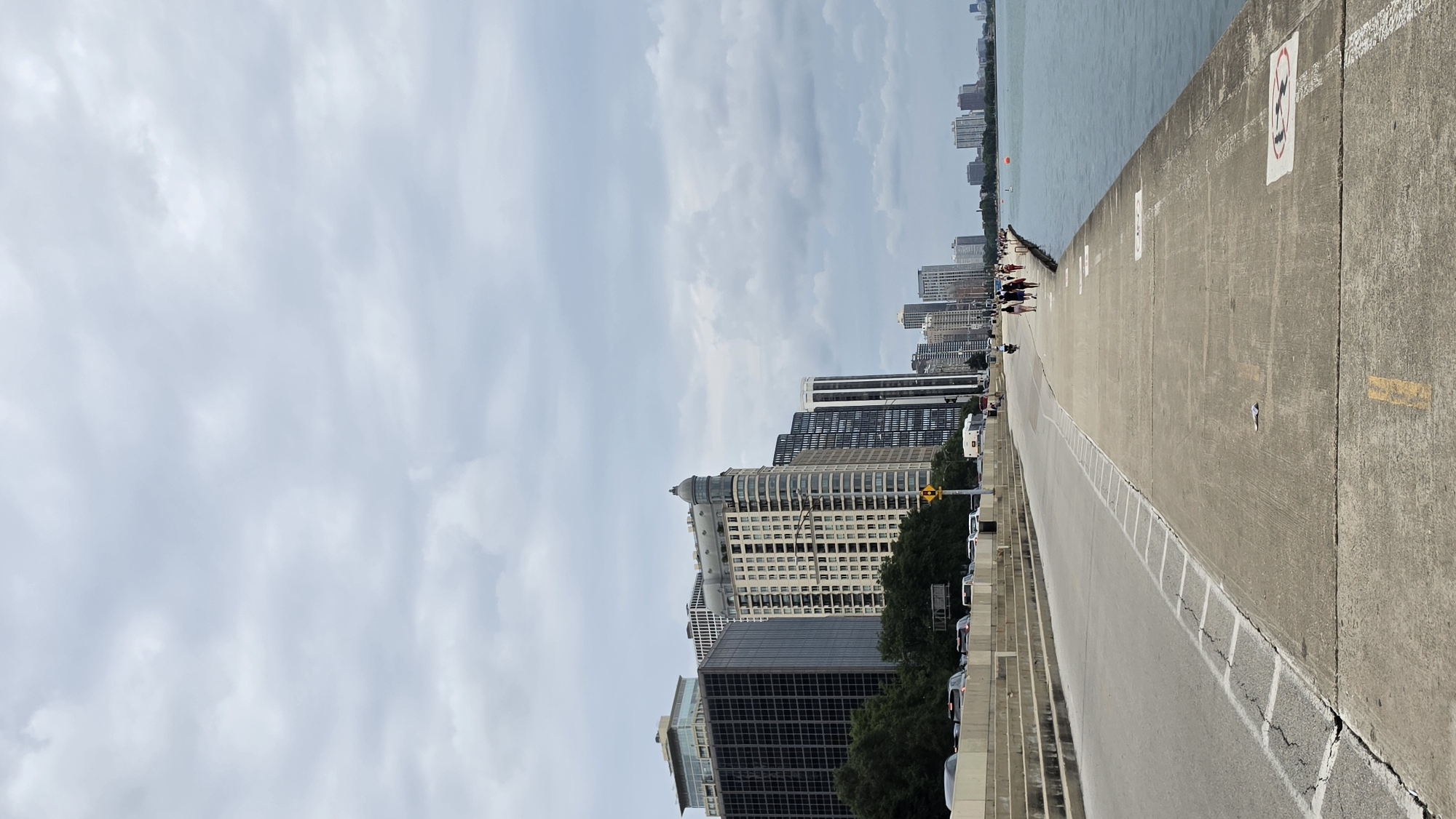 This is a vertical, eye-level, full shot of a cloudy day along the Lake Michigan waterfront in Chicago, USA. The foreground is a wide concrete walkway with painted markings, cracked and worn in places. To the left of the walkway is a paved road with traffic and to the left of the road is a concrete embankment lined with trees and several large buildings. The buildings range in height and architectural style, with one prominent building in the mid-ground featuring a domed top and ornate facade. Further in the distance, more buildings line the waterfront, forming the city skyline. The water of Lake Michigan is on the right side of the image, calm and a muted blue-grey. A few people are walking on the far right of the walkway, their backs to the camera. The sky is overcast with a thick layer of grey clouds.