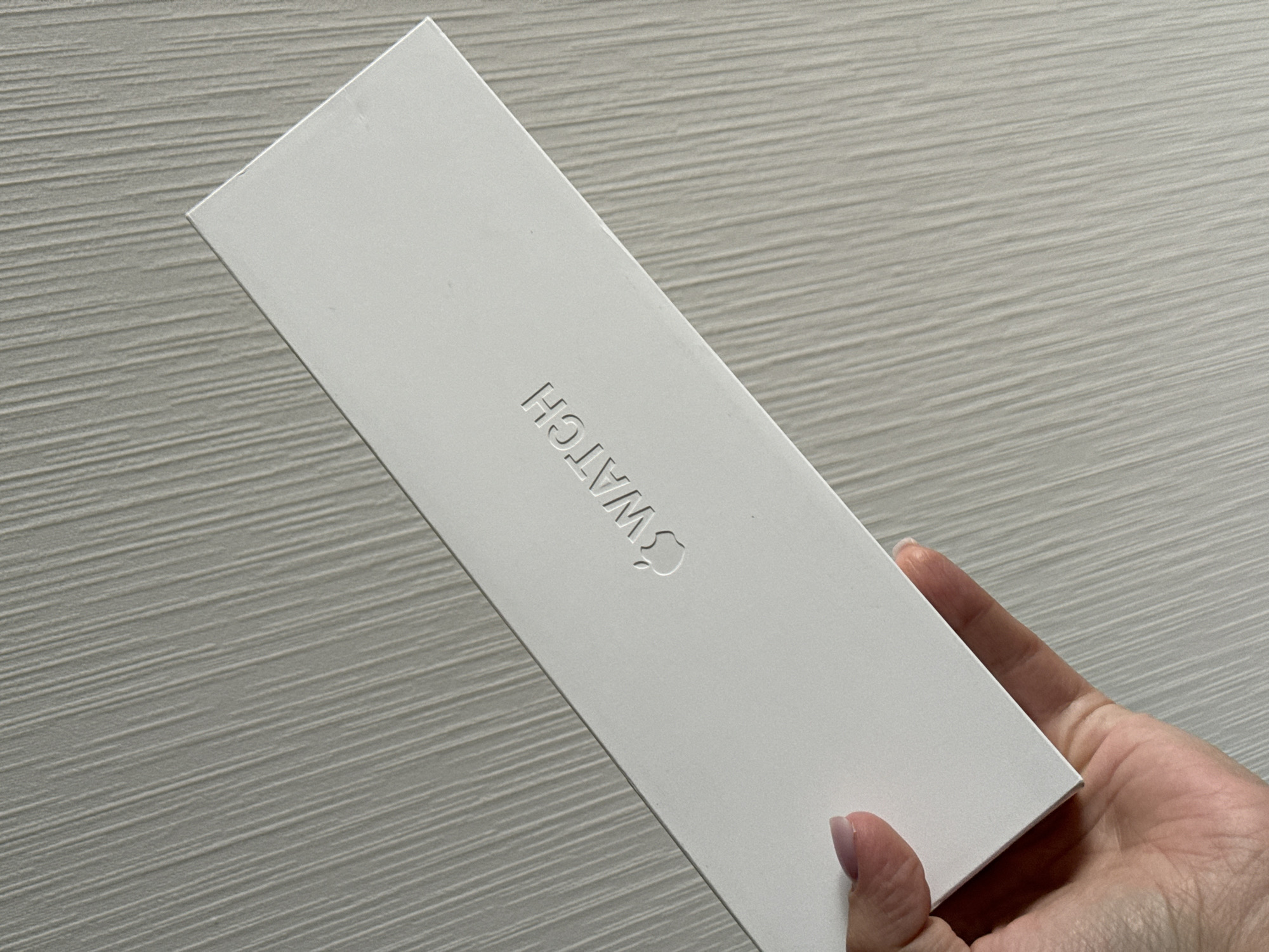 A white box with the embossed logo "SWATCH" is held at an angle by a person's hand. The box is oriented diagonally across the frame, with the bottom left corner closer to the viewer. The background is a textured surface with vertical ridges, appearing to be a wall or similar structure. The lighting suggests an indoor environment, possibly a room. There is no discernible information about the location in Lyubertsy, Russia.