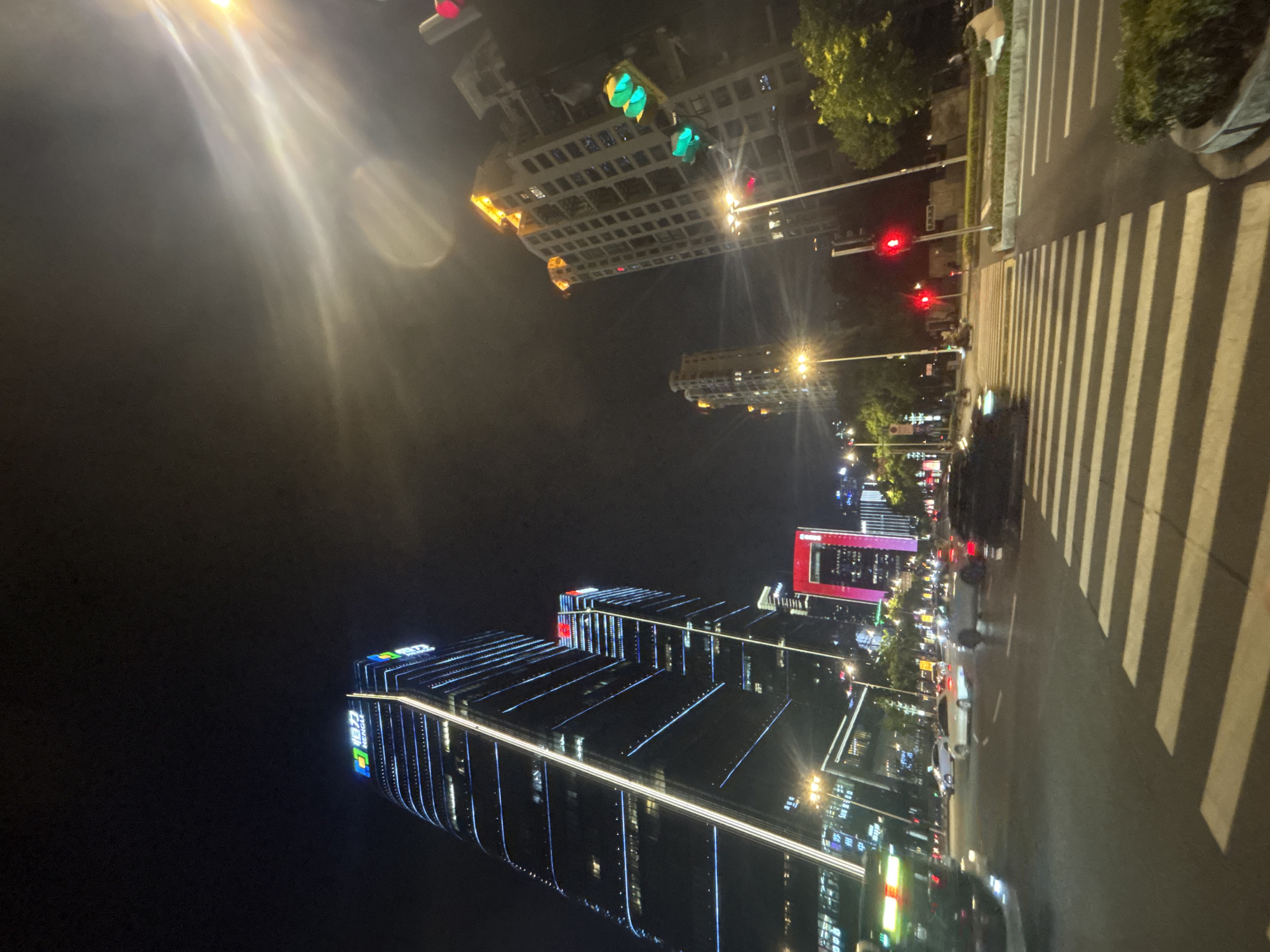 The image shows a street scene at night in Suzhou Industrial Park, China. Modern skyscrapers with illuminated facades dominate the background. A prominent building on the left features a large, brightly lit logo with Chinese characters. Traffic lights are visible, some showing red, indicating a controlled intersection. Several cars are on the road, some appearing to be stopped at the intersection, while others are in motion. A pedestrian crossing is clearly marked on the asphalt. The sky is dark, with some lights from buildings and streetlights casting illumination on the scene. Trees and landscaping are present along the sides of the road, adding a touch of nature to the urban environment. The overall atmosphere is one of a bustling, modern city at night.
