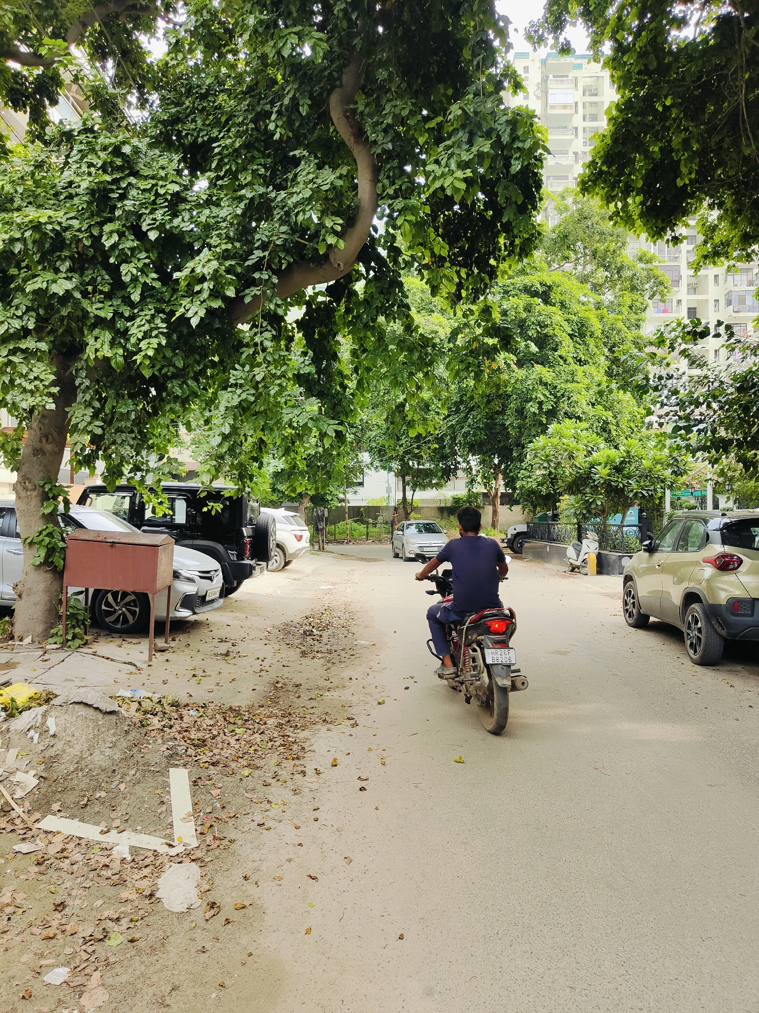 A man is riding a motorcycle down a street lined with trees in Gurgaon, India. He is wearing a dark blue t-shirt and dark pants. The motorcycle has a red tail light and a license plate that reads "HR26F 88206". On the left side of the street, there are several parked cars, including a black SUV and a white car. There is also a trash can on the left side of the street. In the background, there are tall buildings, indicating an urban environment. The street is paved and covered with some fallen leaves. The scene appears to be during the day, with overcast sky.