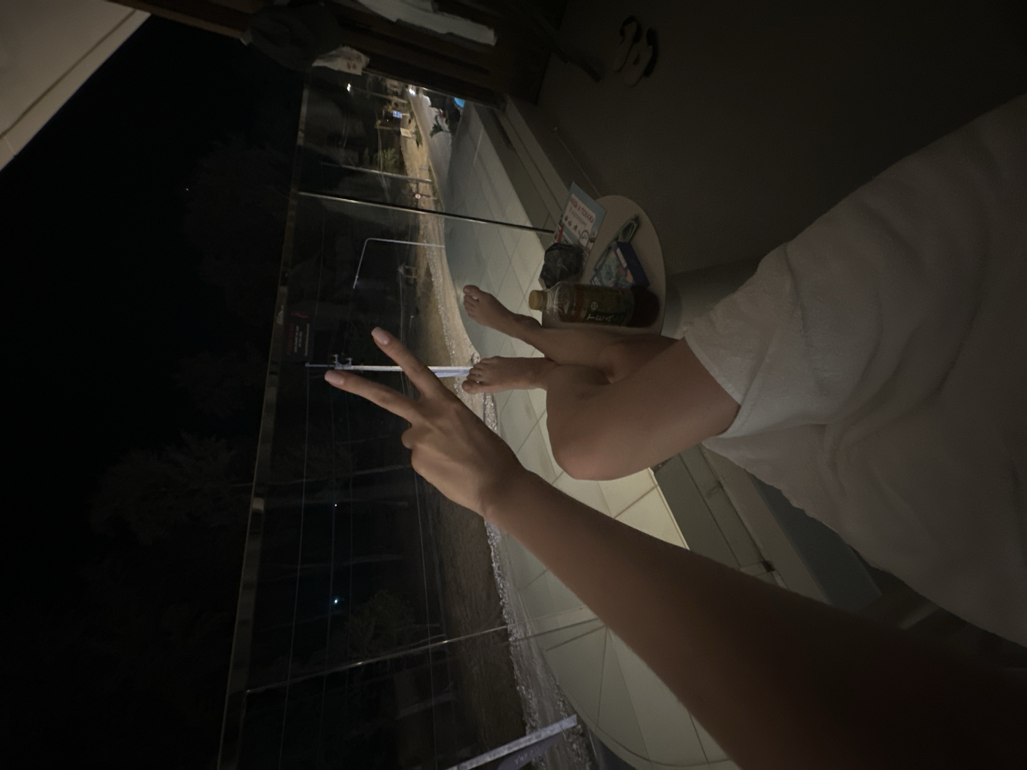 The image displays a nighttime scene from a balcony in Choeng Thale, Thailand. A person is seated with their legs extended and bare feet visible, partially covered by a white towel or garment. Their right arm is raised, with the hand forming a "peace" sign (V-sign) with two fingers extended. Adjacent to the person's legs, on the right, is a small, round, light-colored table. On this table are a plastic bottle containing a brown liquid, a dark object, a magazine or book featuring Thai text, and a blue pack of cigarettes.
The balcony features a clear glass railing with a metal top bar, offering a view of the street below. The street is paved and appears to be a two-lane road, illuminated by several streetlights. Vehicles, including one with headlights on and another parked, are discernible on the street. Utility poles and various signs with Thai script are visible along the roadside. The background consists of dark trees silhouetted against the night sky. On the right side of the balcony floor, near a dark wooden doorframe, a pair of light-colored flip-flops are present, along with a piece of light-colored fabric hanging near the doorframe.