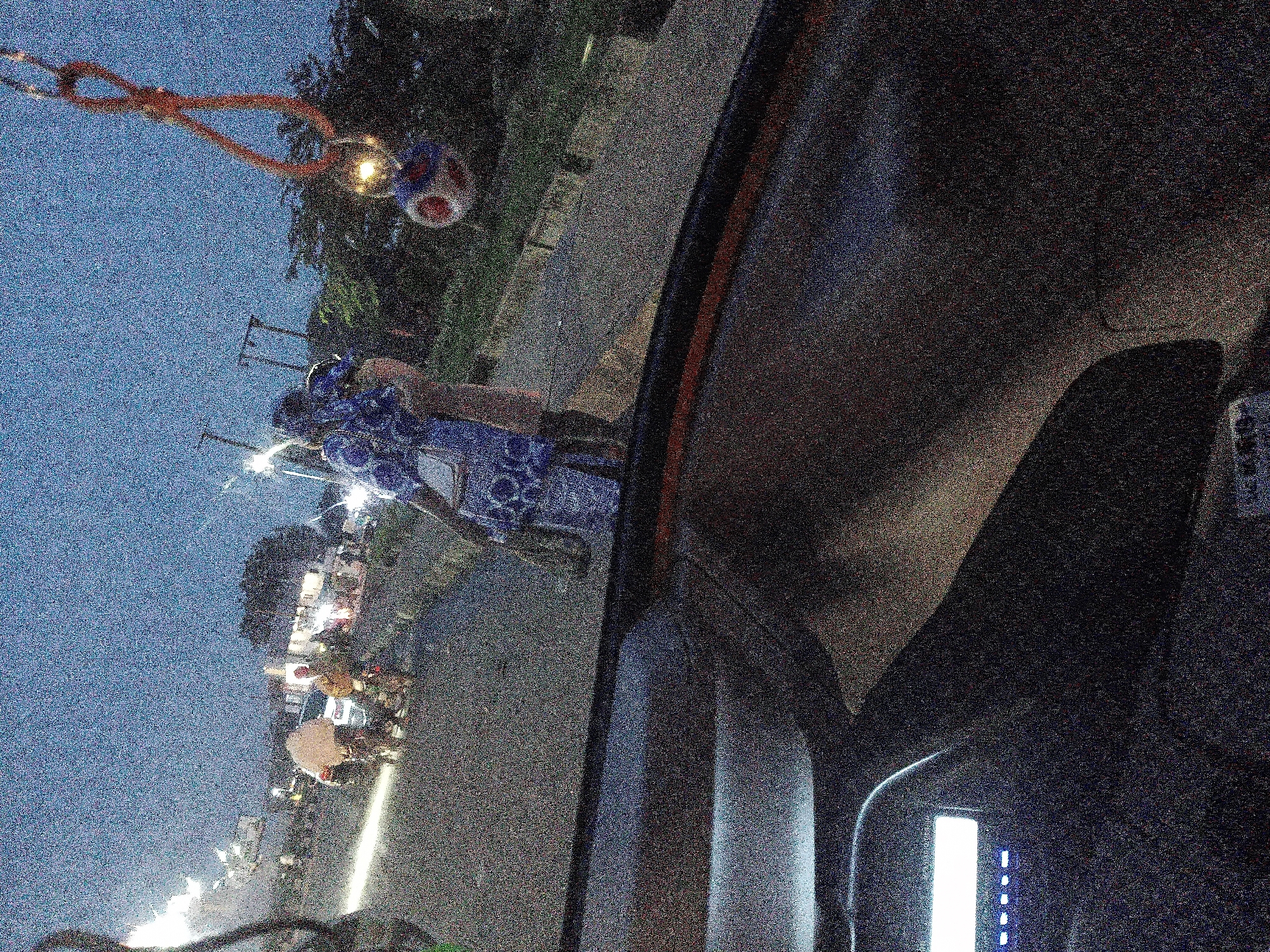 The image depicts a nighttime scene at Barakin Lerawi, Nigeria, likely on a street. In the foreground, the dark interior of a vehicle is visible, with a glowing digital display on the left. The middle ground features two individuals embracing; one person is wearing a blue and white patterned dress and appears to be holding a white handbag, while the other person is wearing a lighter colored garment. Behind them, several motorcycles and people are present, with some motorcycles parked and others in use, illuminated by streetlights and vehicle headlights. The overall atmosphere is dimly lit, suggesting dusk or nighttime, with a grainy texture throughout the image, common in low-light photography. The background shows trees and what appear to be structures, with overhead power lines also visible. A decorative object, possibly a keychain, hangs from a string in the upper right corner of the frame.